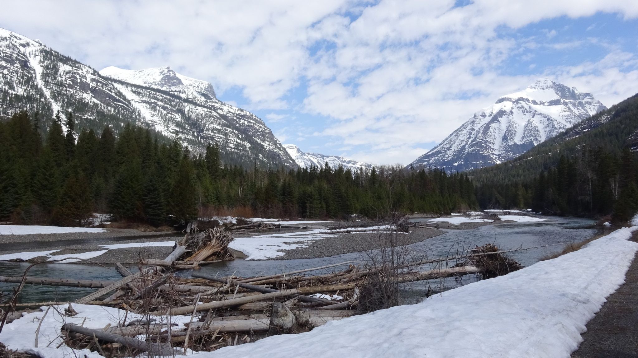 A wide river with snow-covered banks and driftwood, surrounded by snowy mountains. - Upper McDonald Creek Trail
