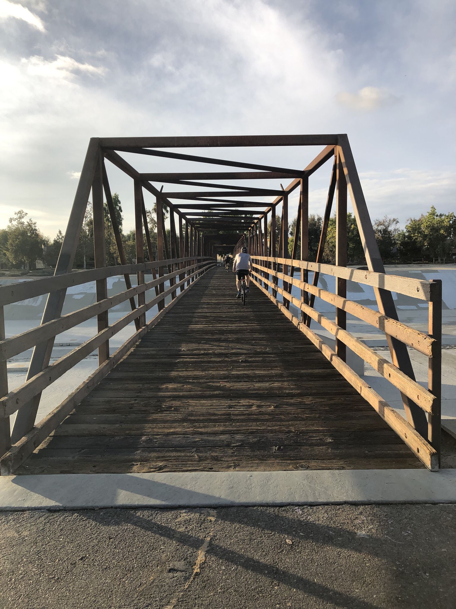 Cyclist on a long, dark metal and wooden bridge under a bright, cloudy sky. - Santa Ana River Trail