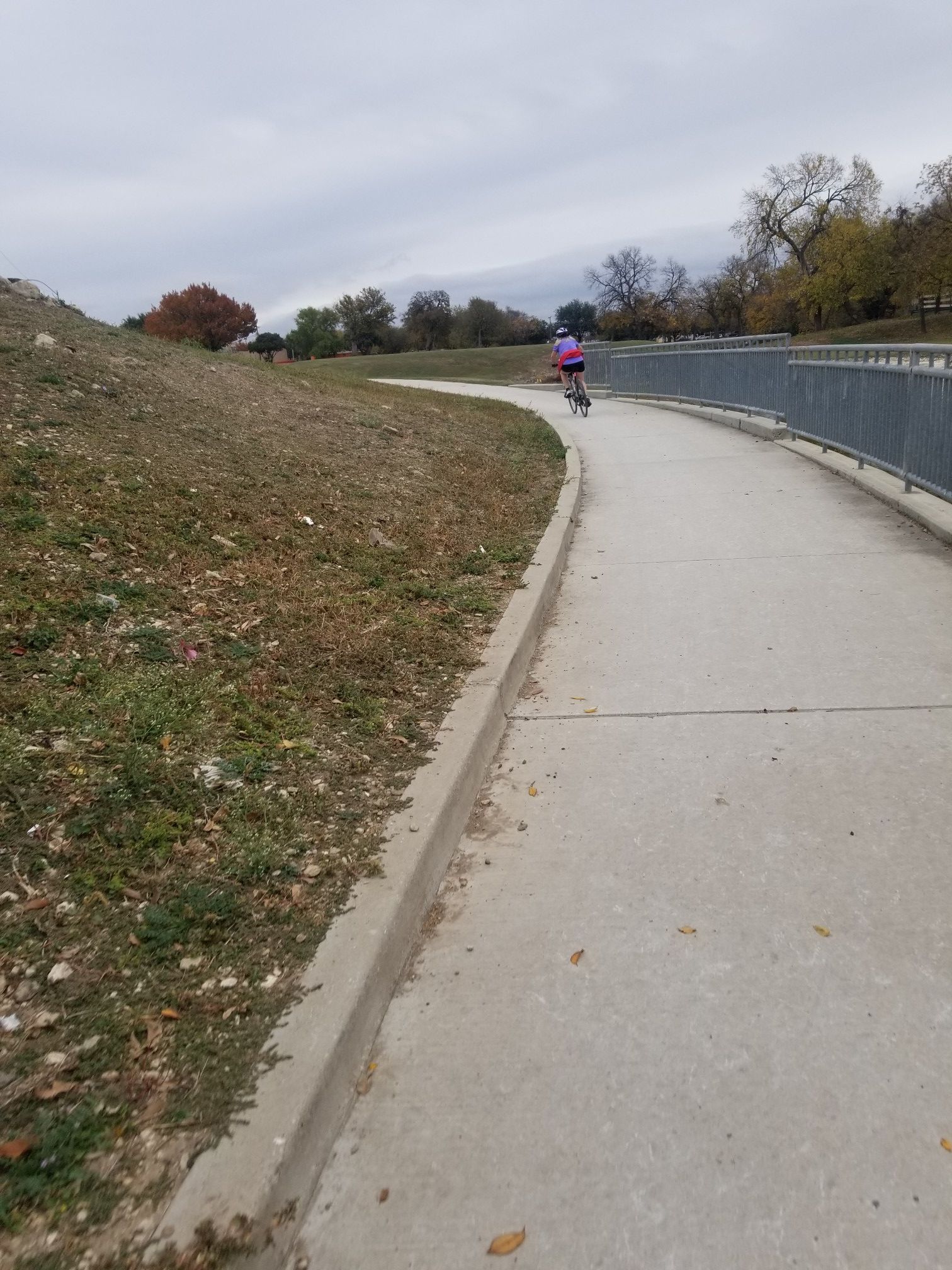 A person cycles on a paved path next to a grassy slope under a cloudy sky. - Trinity Trails: Fort Worth Nature Center Loop