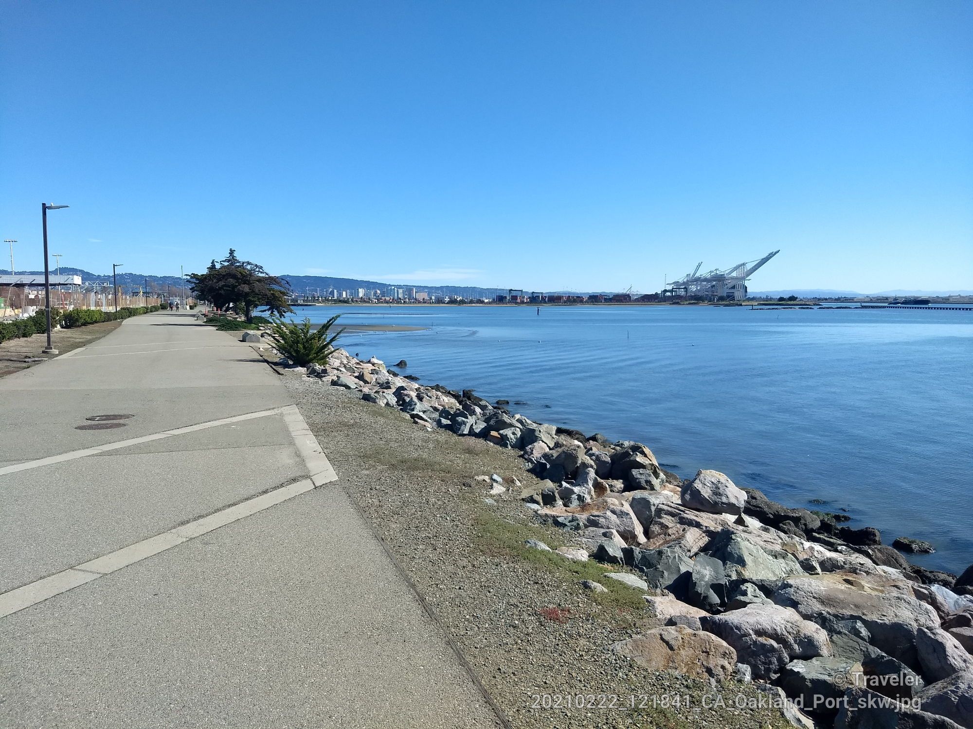 Paved path along a rocky shore, calm water, and distant industrial cranes. - Downtown Berkeley to Middle Harbor Shoreline Park loop