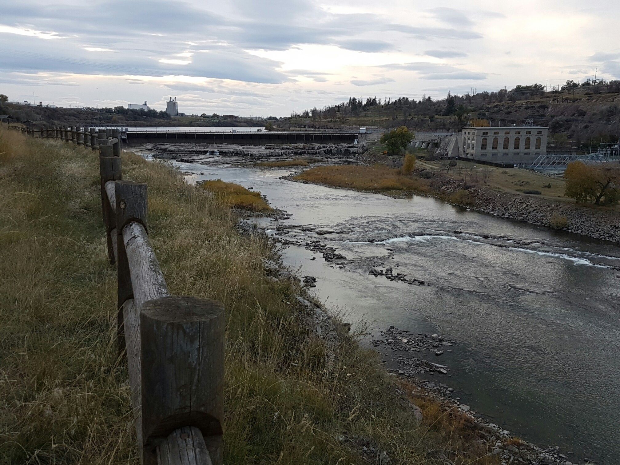 Overcast view of a river flowing past a wooden fence and industrial buildings. - River's Edge Trail