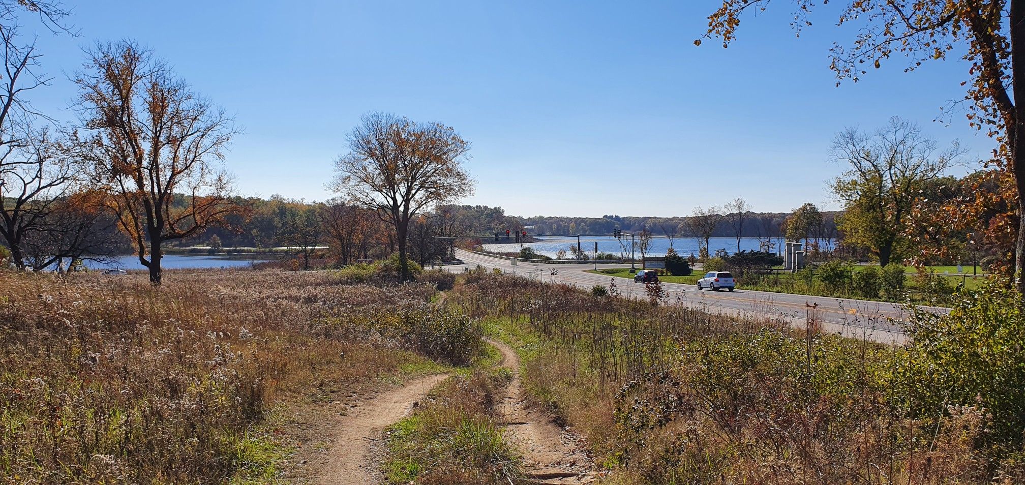 A wide view of a lake, road, and dirt path on a clear autumn day. - View of Tomahawk Slough – Tomahawk Slough loop from Worth