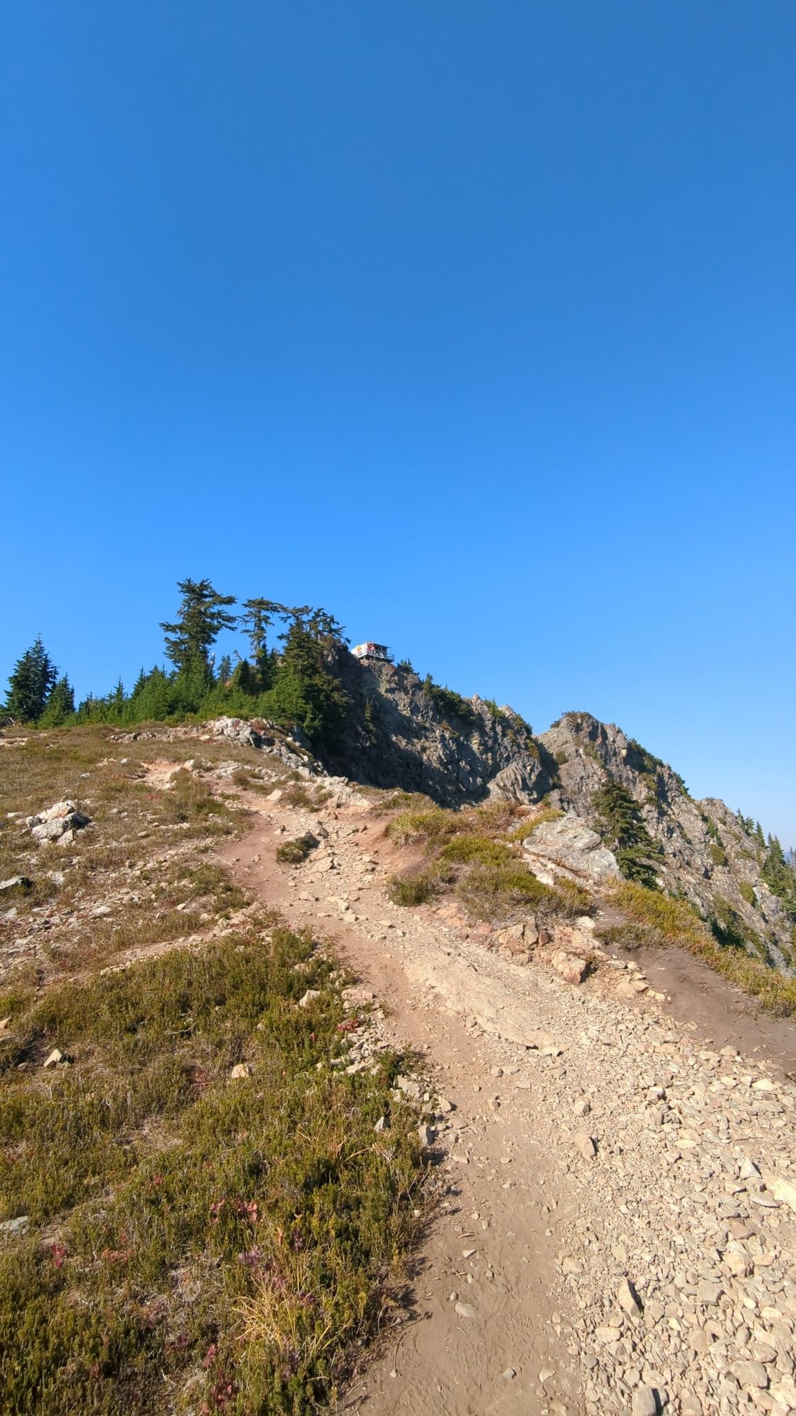 Dirt trail ascending a rocky, grassy slope towards a distant mountain lookout. - Park Butte Lookout – Mazama Park shelter loop from Ridley Creek Trailhead