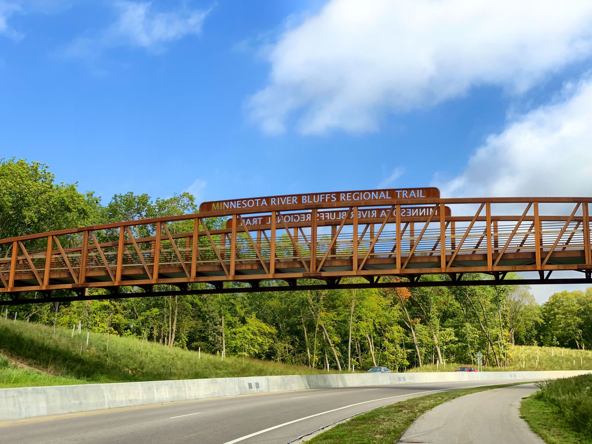A metal pedestrian bridge crosses over a highway with green trees. - Bluff Creek Drive trail access – Seminary Fen SNA loop from Bavaria Rd