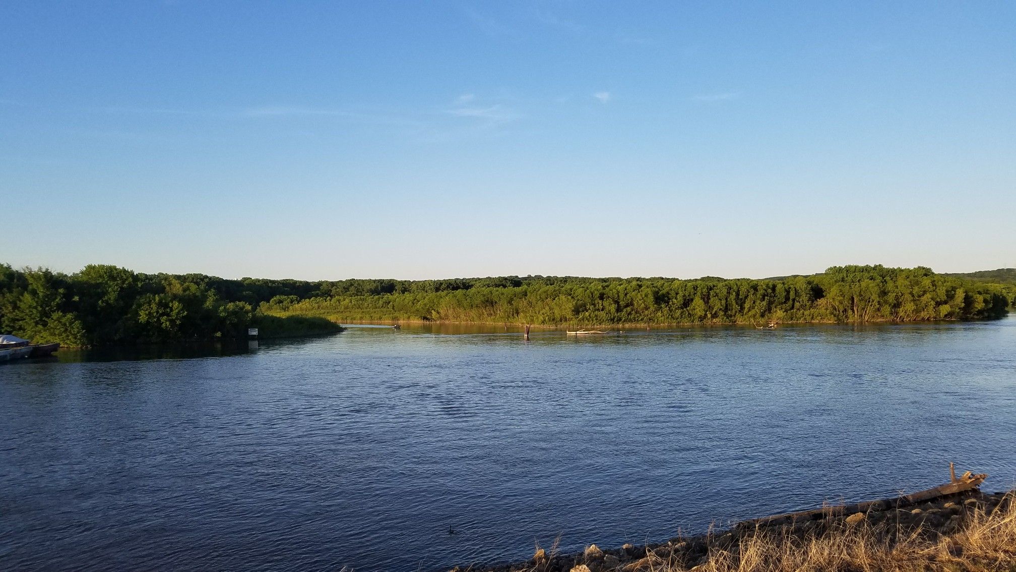 Wide river flowing past a distant green forested bank under a clear sky. - Mississippi River Greenway