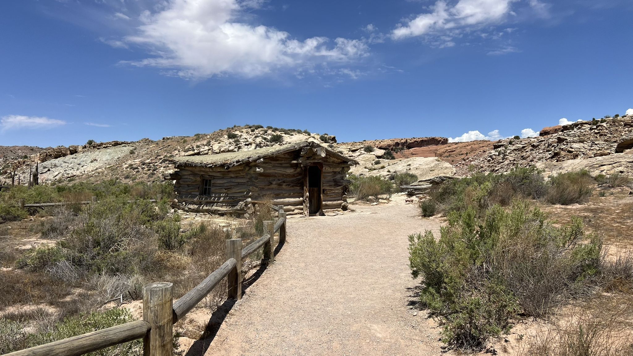 A rustic log cabin next to a dirt path and fence under a blue sky. - Petroglyph Trail