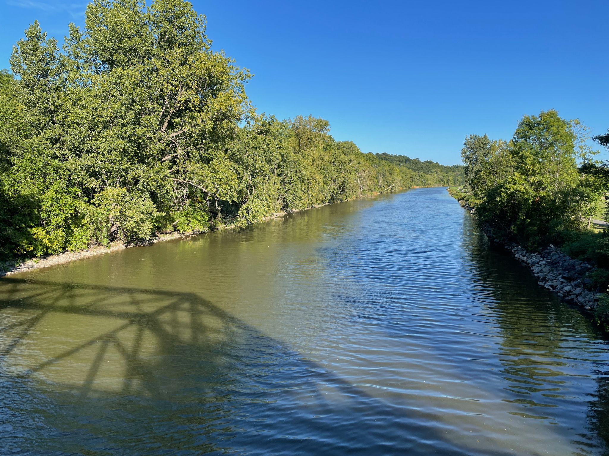 A wide river flows between tree-lined banks under a clear blue sky. - Macedon Bile Bridge – Erie Canalway Trail loop from Lyons