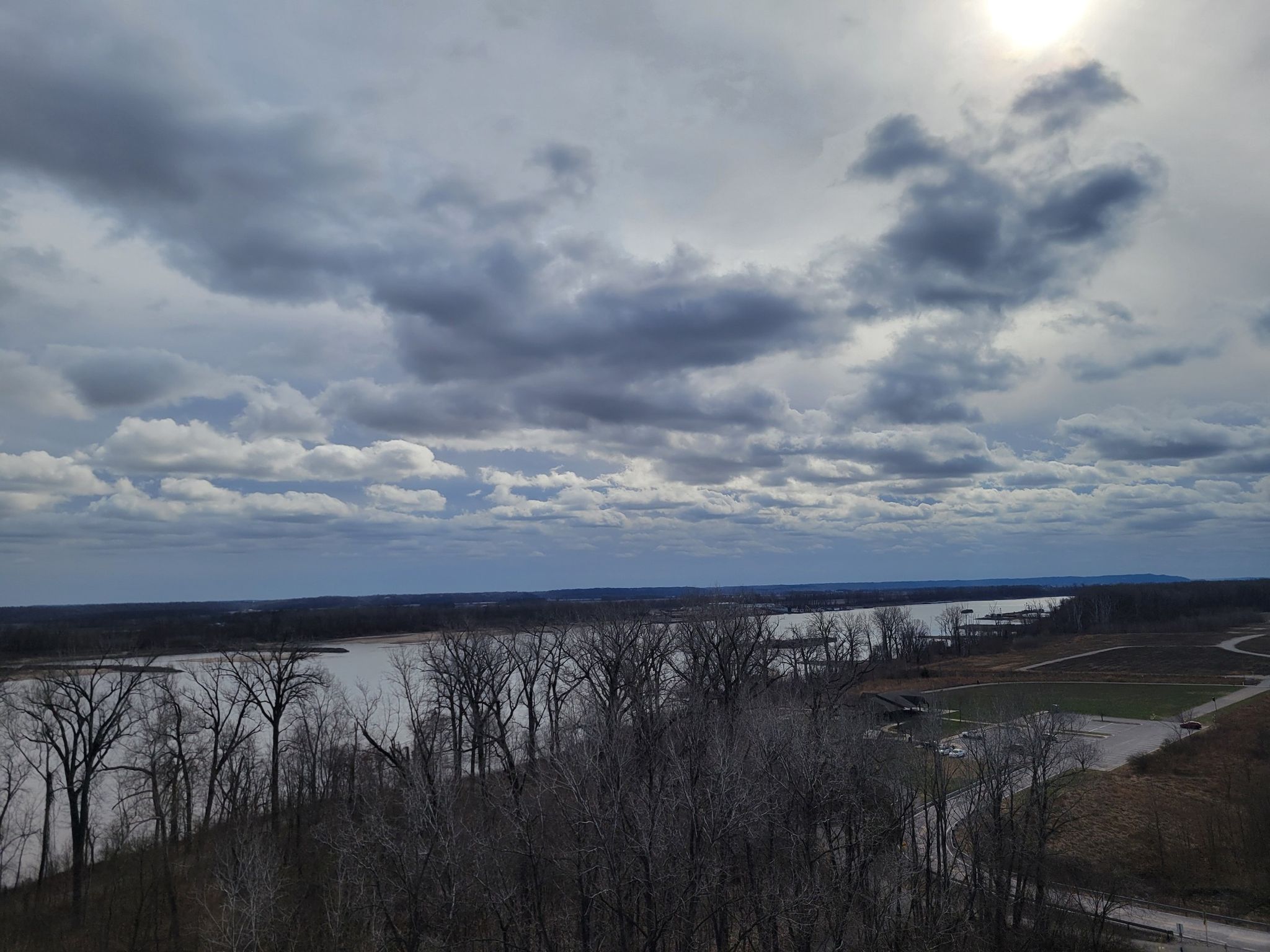 Wide river with bare trees under a cloudy sky, sun peeking through. - Cliff Cave County Park Loop