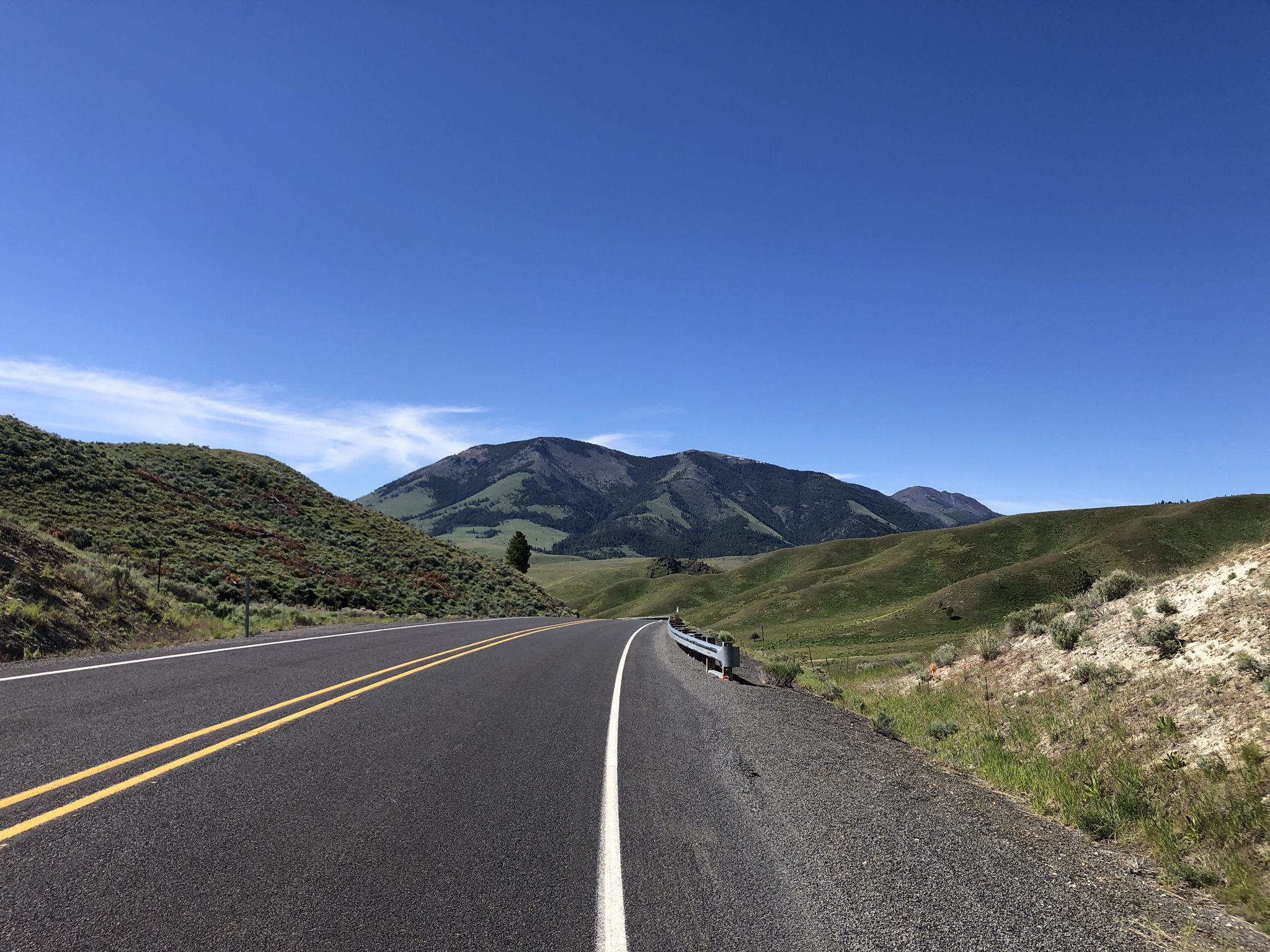 Paved road curves right through green hills toward distant mountains under blue sky. - Eldorado Pass via US26