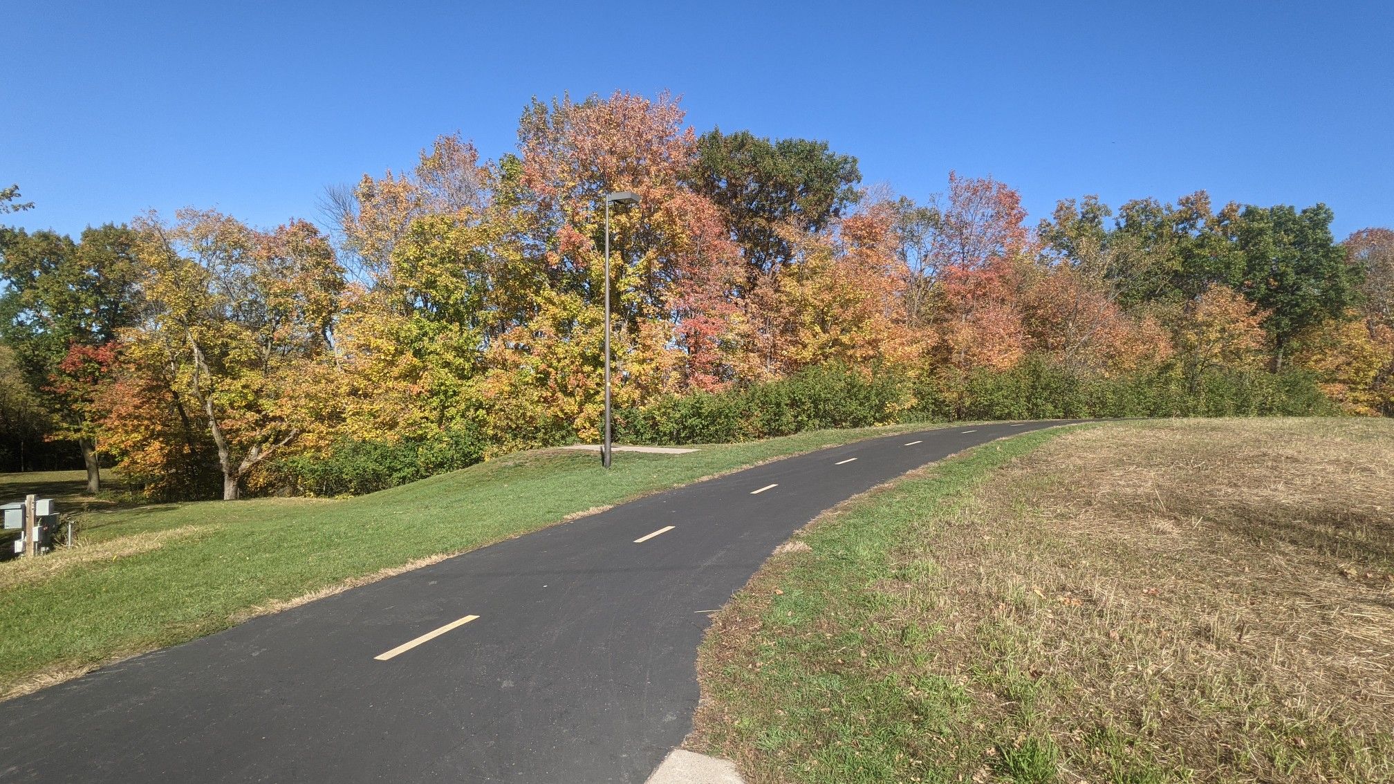 Paved path curving past vibrant autumn trees under a clear blue sky. - Elm Creek Park Trailhead – Elm Creek Park Reserve loop from 96 Av N