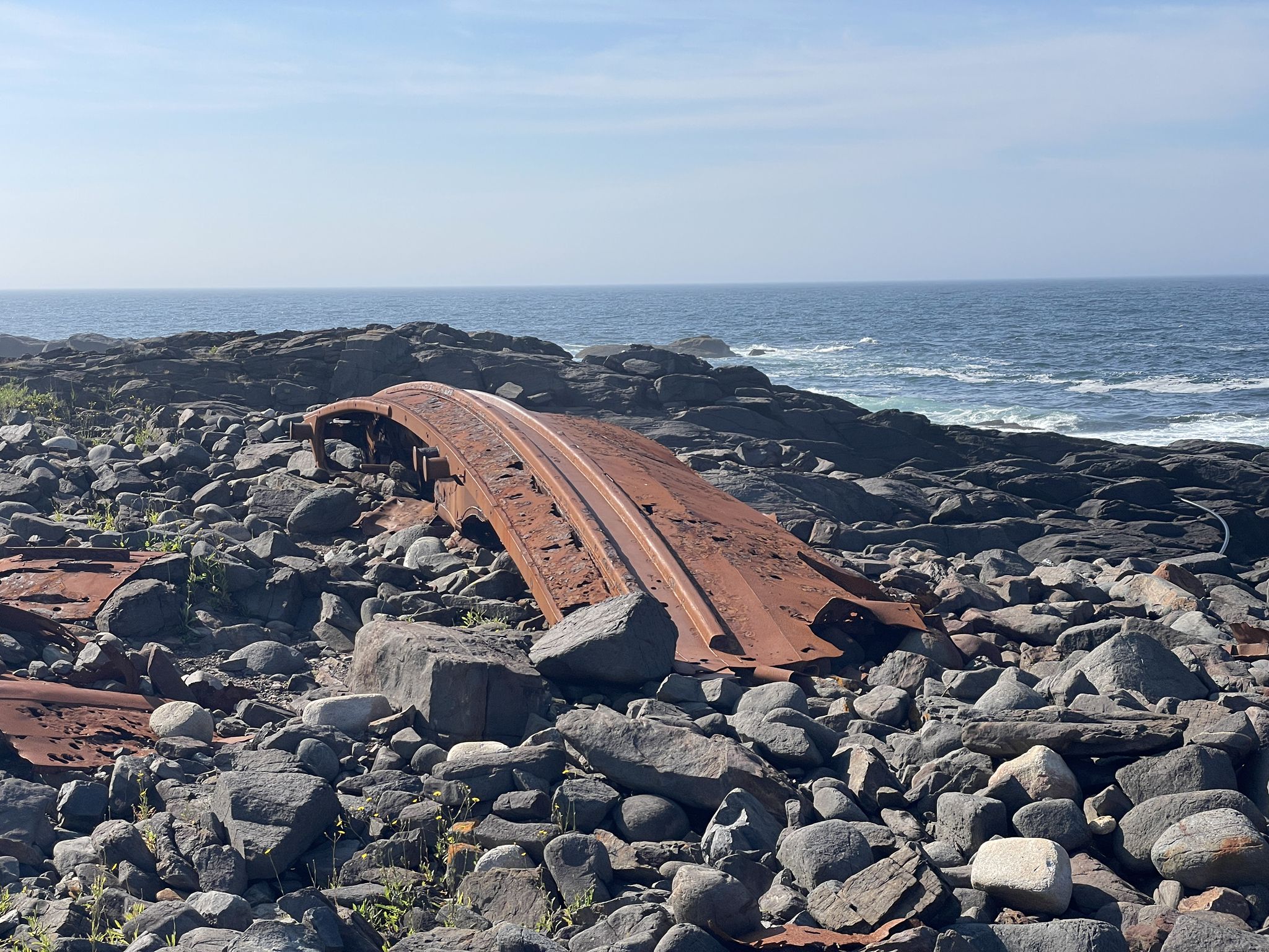 Rusty metal wreckage on a rocky shore with ocean under clear sky. - Cliff Trail
