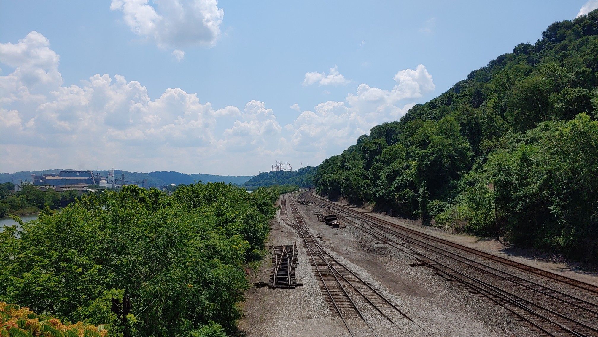 Multiple train tracks run alongside a forested hill under a cloudy sky. - Red Waterfall – Homestead Pump House loop from Homestead