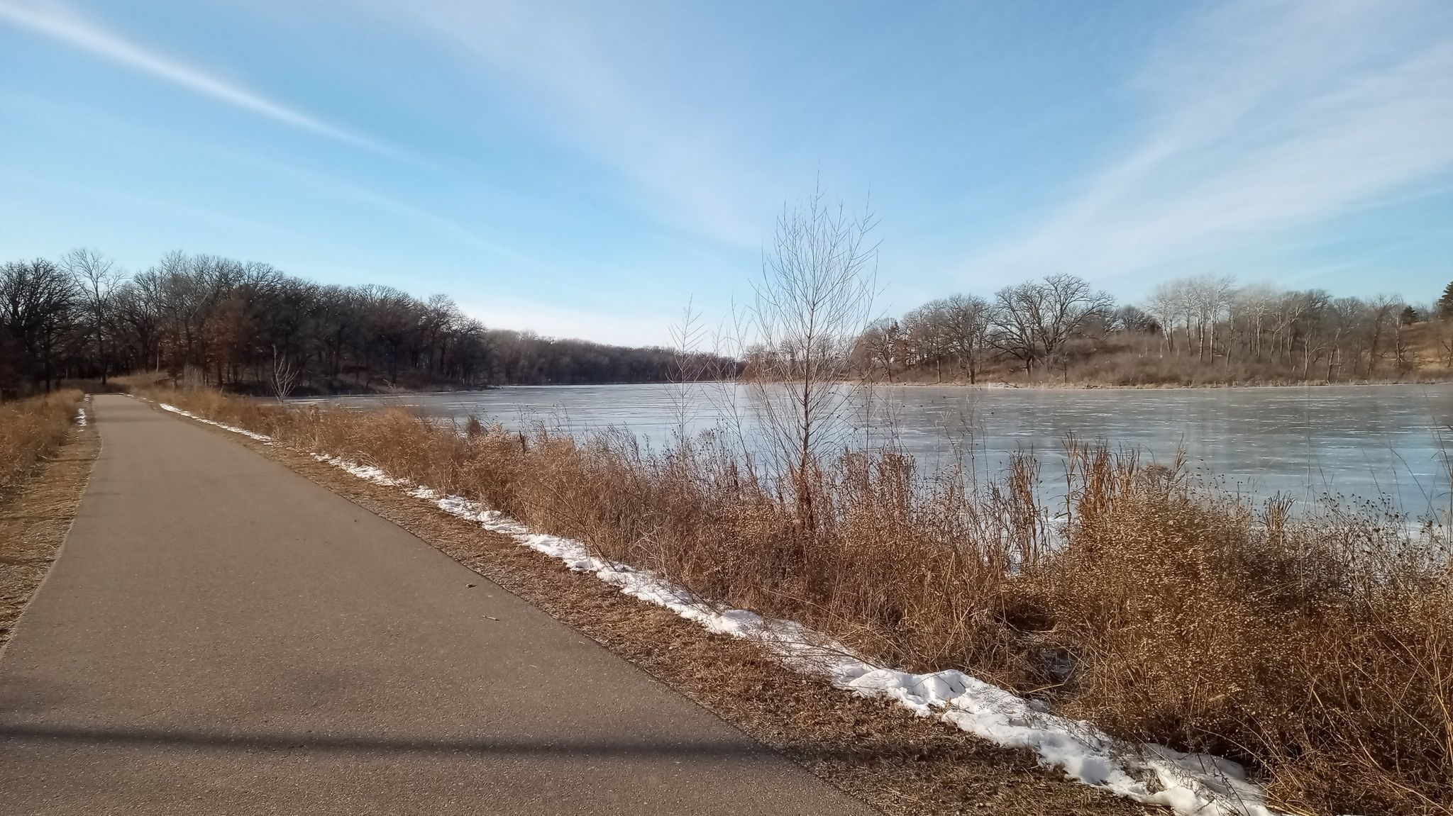 Paved path alongside a partially frozen river with bare trees under a bright sky. - Whitetail Woods Southern Loop