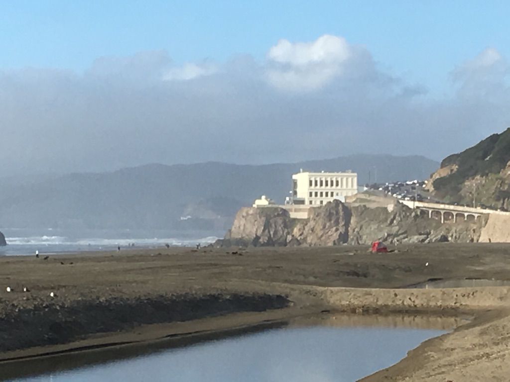 Hazy view of a building on a cliff, sandy beach, and calm water channel. - Murphy Windmill – Golden Gate Park loop from Judah Street & 9th Avenue