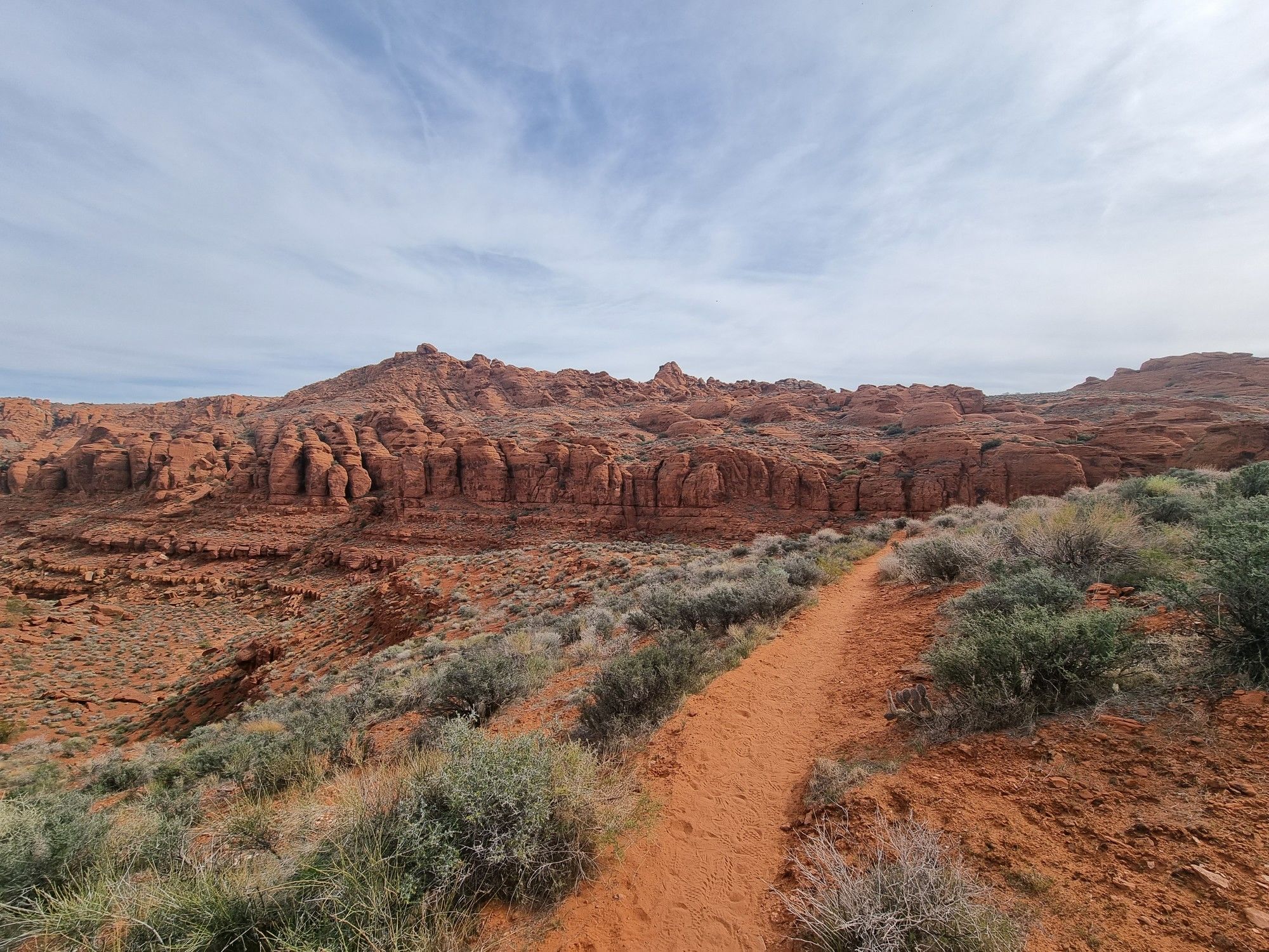 A dirt trail winds along a hillside overlooking red rock formations under a cloudy sky. - Paradise Rim and Scout Cave Loop