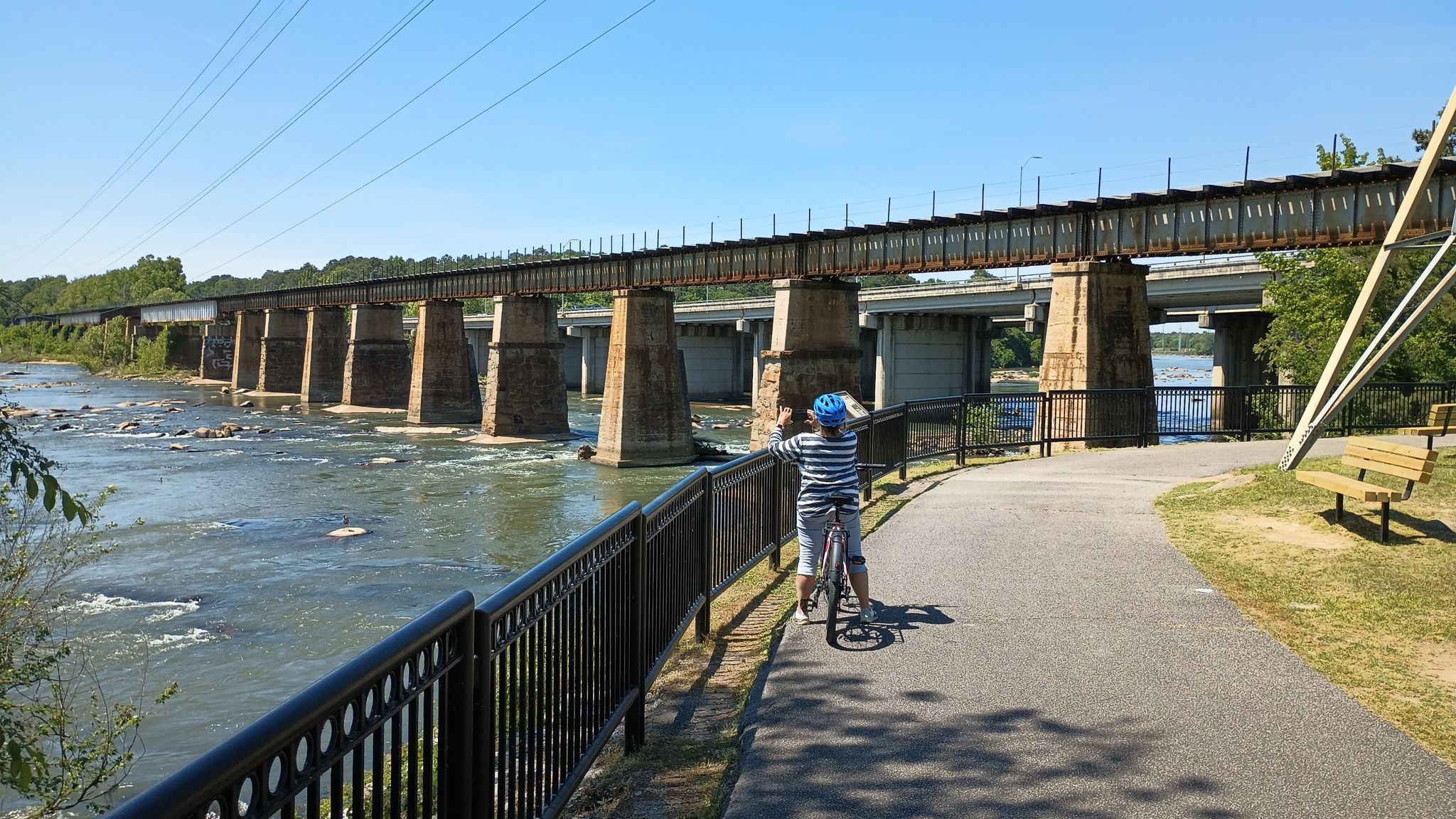 Child on a bicycle by a river, looking at a long bridge under clear sky. - Three Rivers Greenway