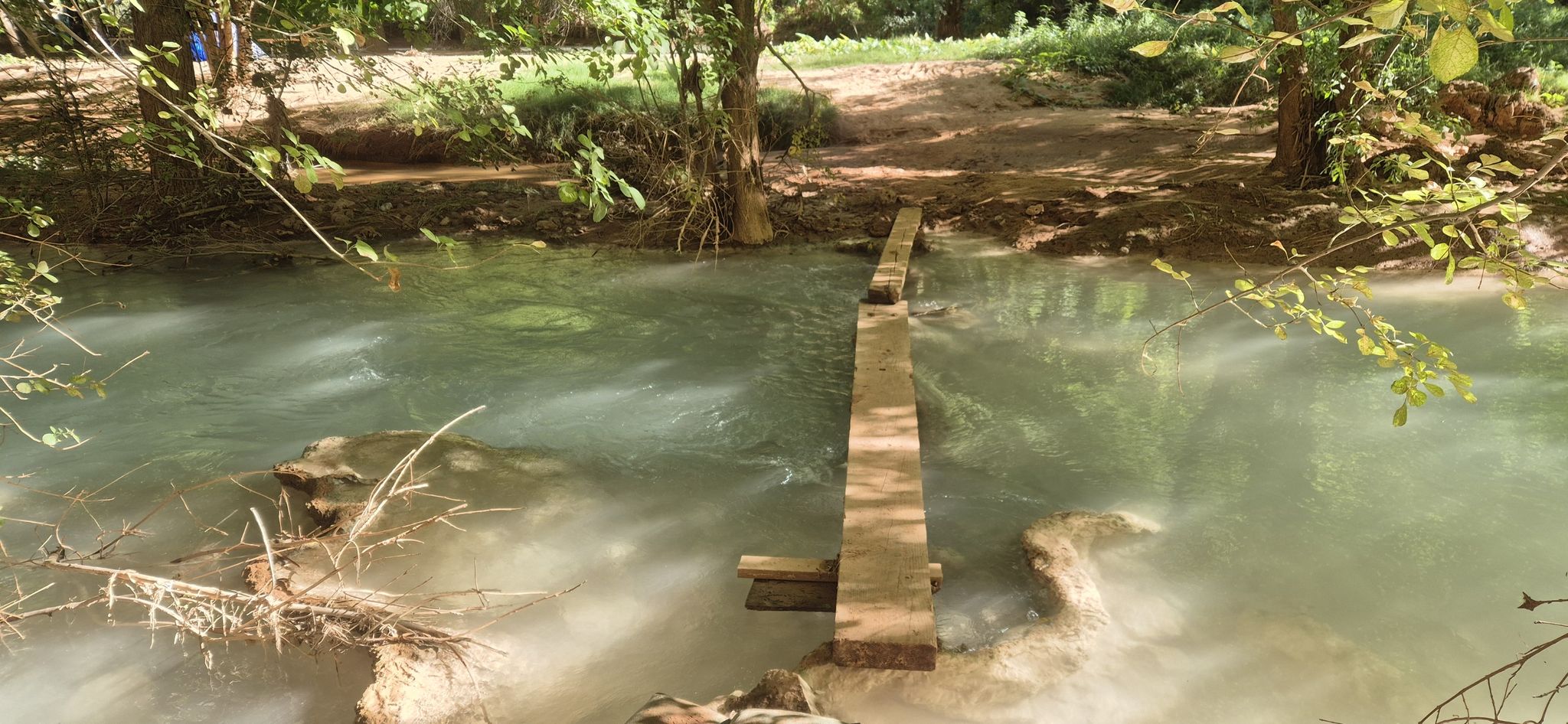Wooden planks form a makeshift bridge over a shallow, milky turquoise river. - Mooney Falls loop from Havasu Falls