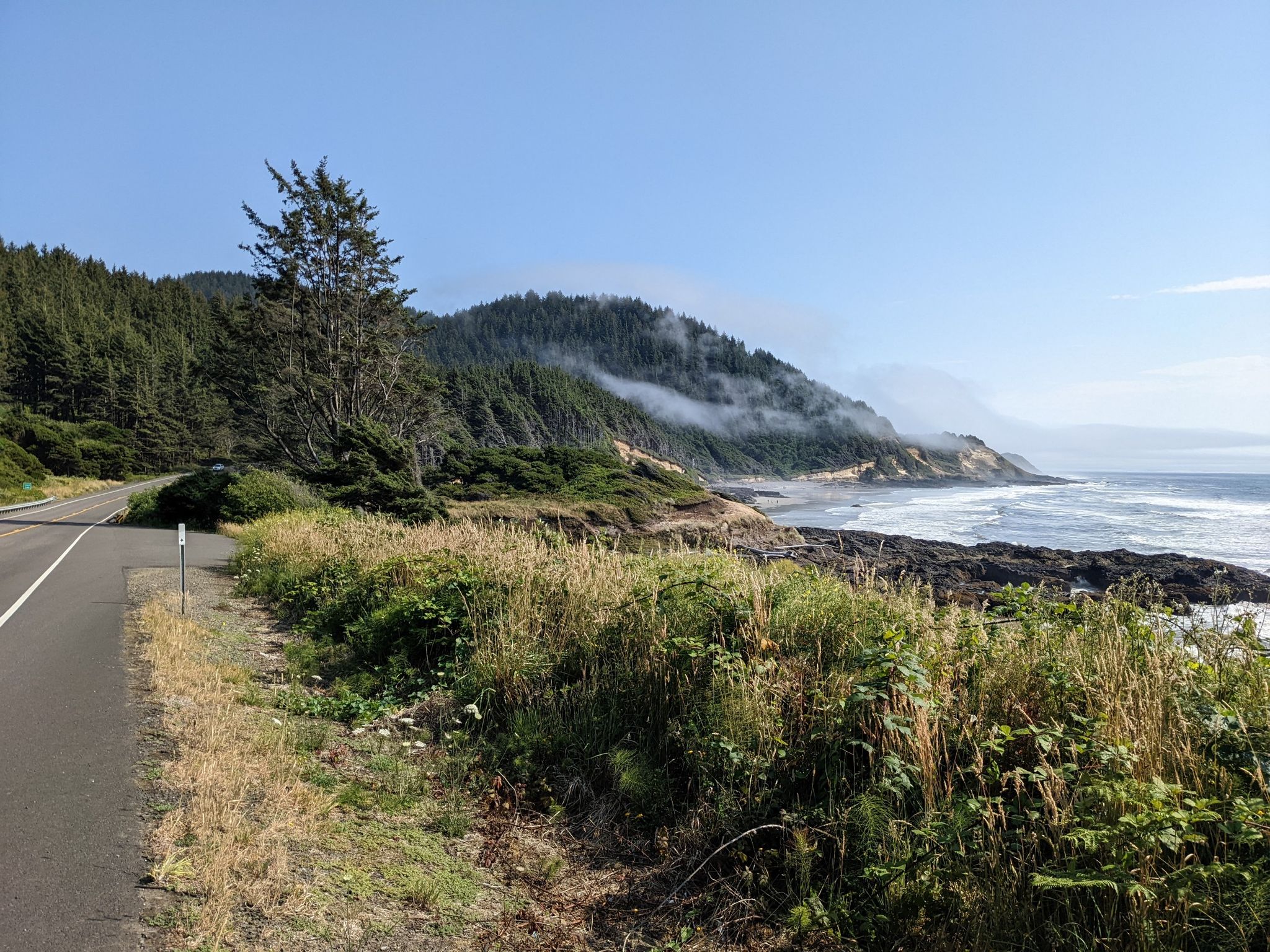 Coastal road next to a rocky shore with waves and misty forested hills. - Oregon Coast Scenic Bikeway