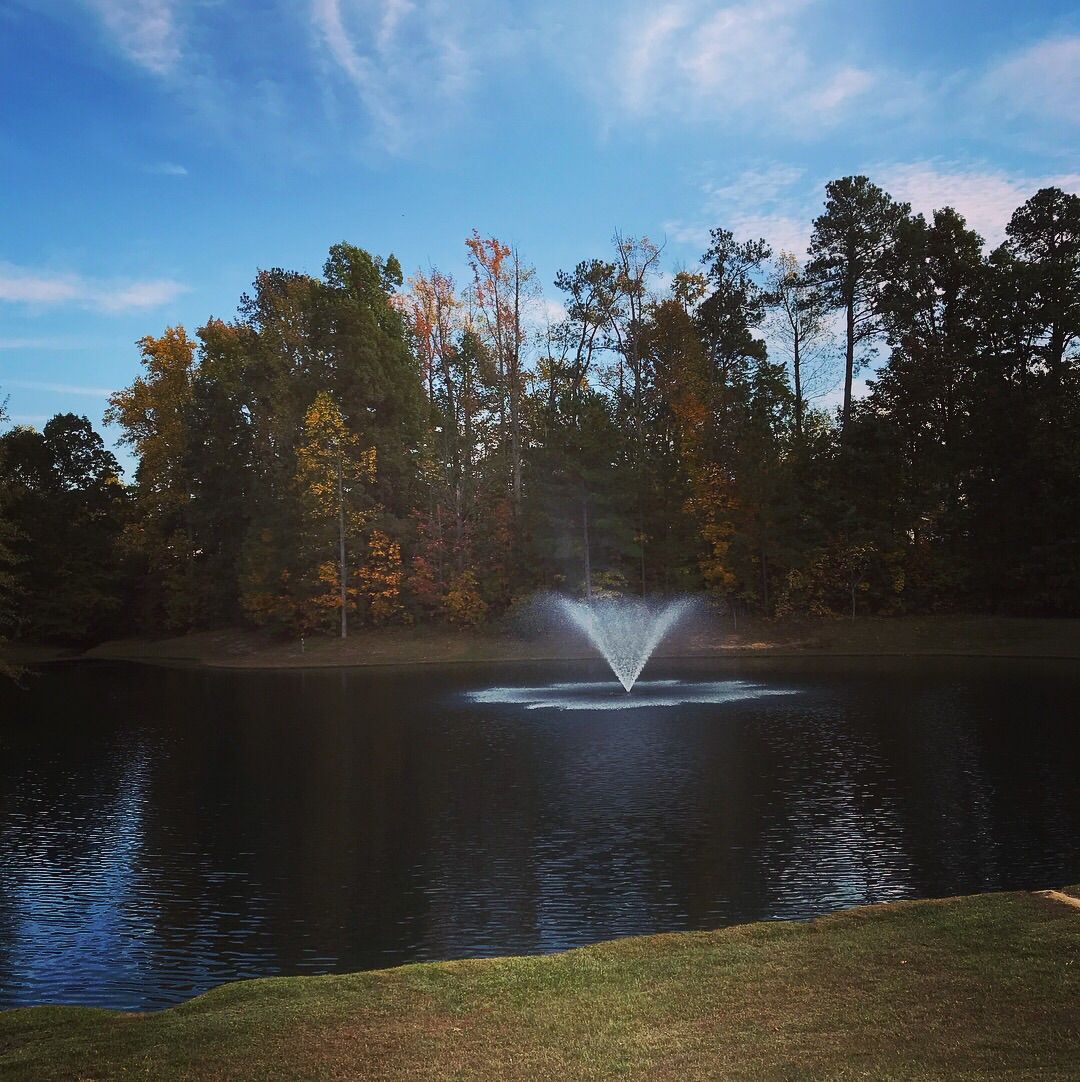 A fountain sprays water in a pond surrounded by colorful autumn trees. - Park Village Greenway