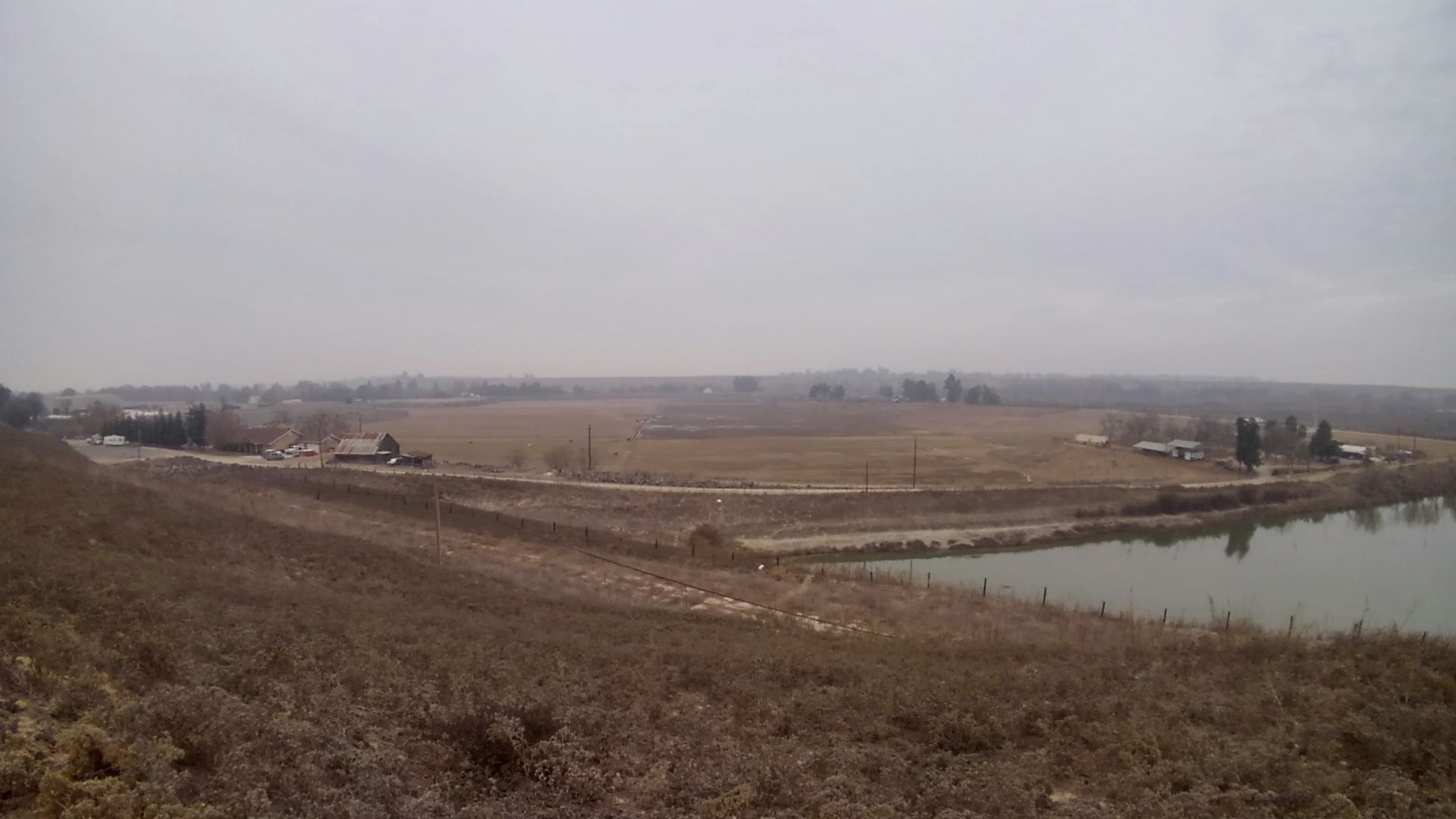 An overcast view of a river, fields, and distant buildings. - Woodward Park – Woodward Park loop from Parkway Trust / Caglia