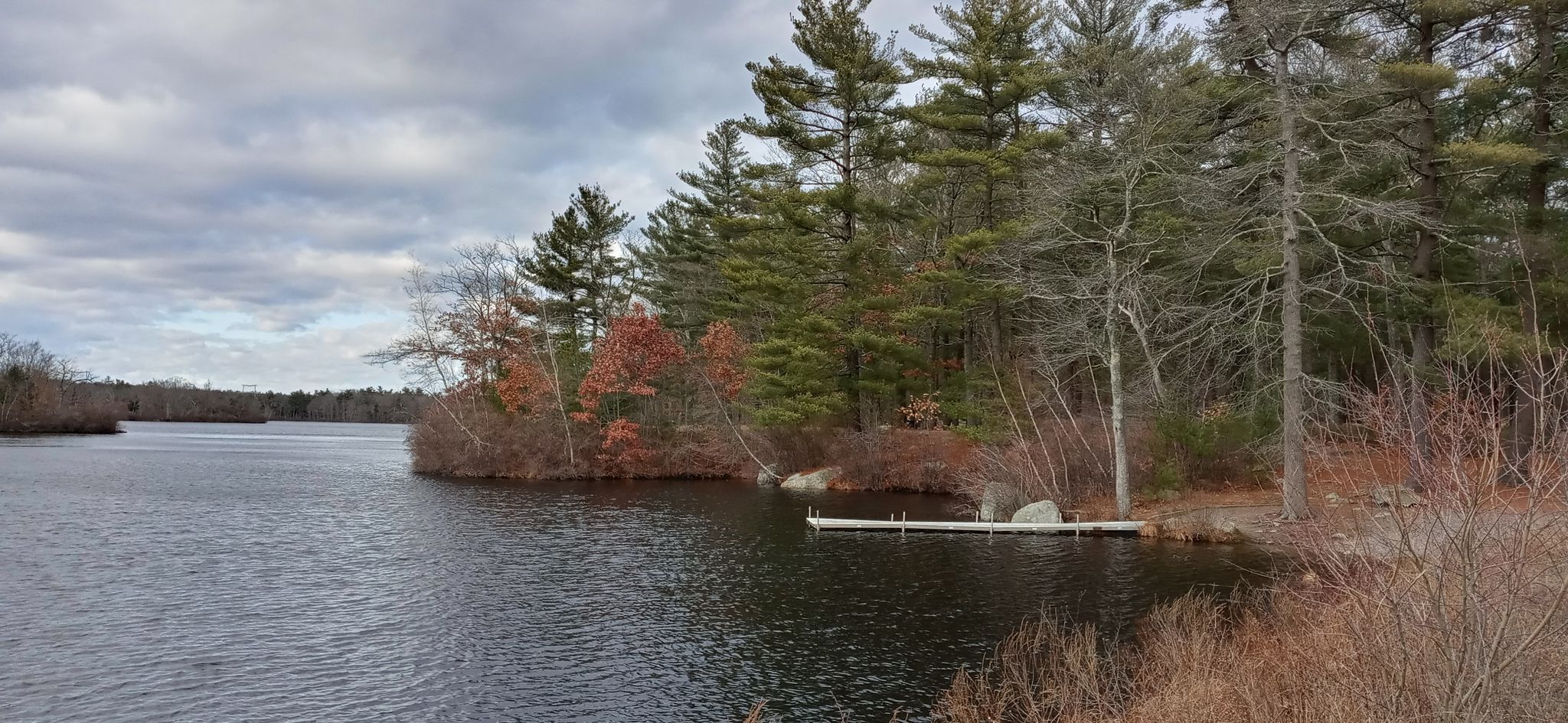 Dark lake water with a small dock, surrounded by trees under a cloudy sky. - Ames Nowell Loop