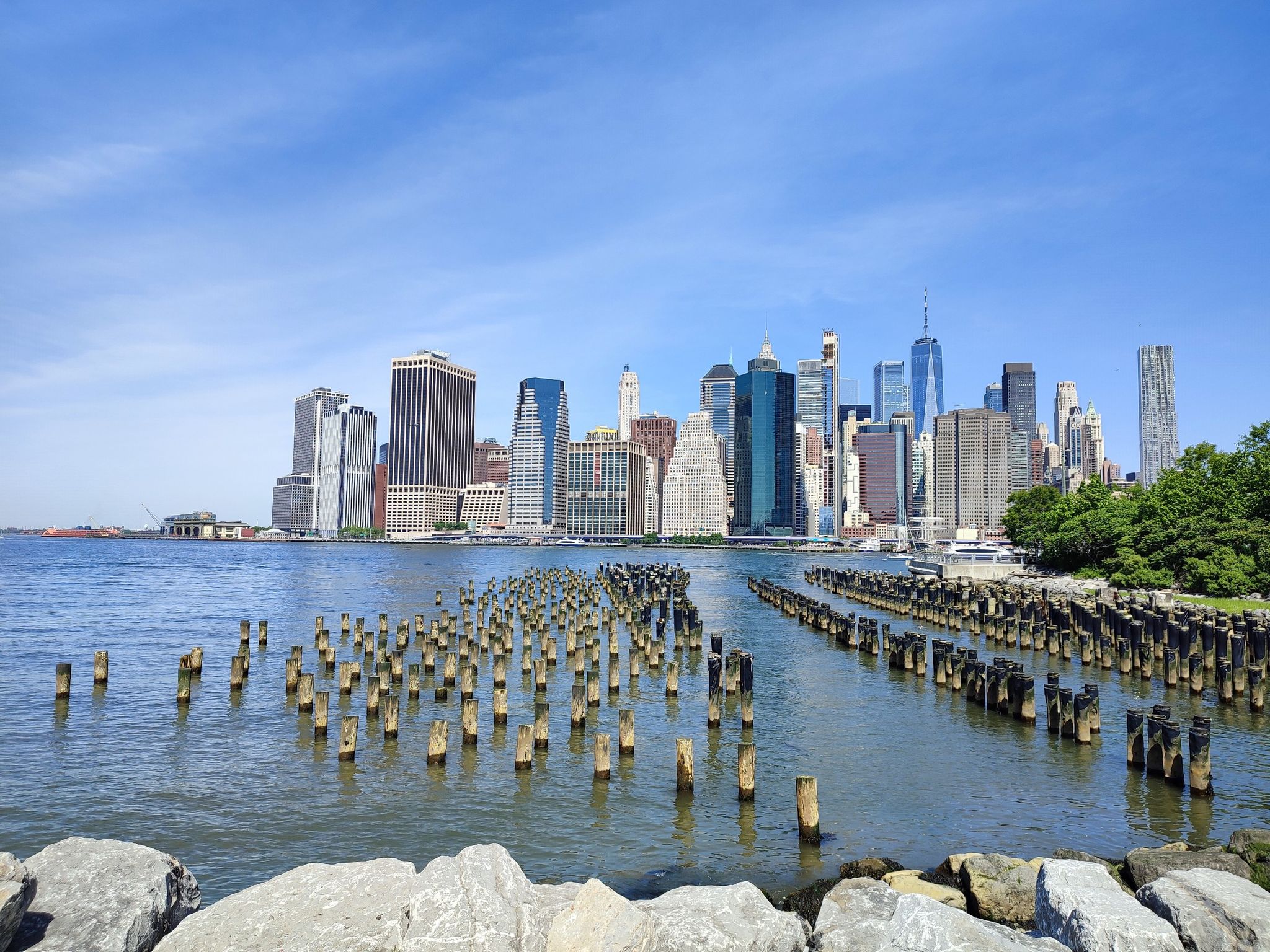 City skyline across water with many old pier pilings under a clear blue sky. - One World Trade Center – East Coast Memorial loop from Wall Street