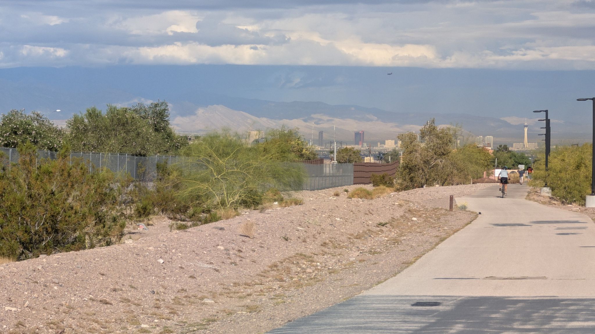 Cyclist on paved path through desert, distant city under cloudy sky. - Railroad Pass Hotel & Casino – Acacia Park loop from Green Valley High School