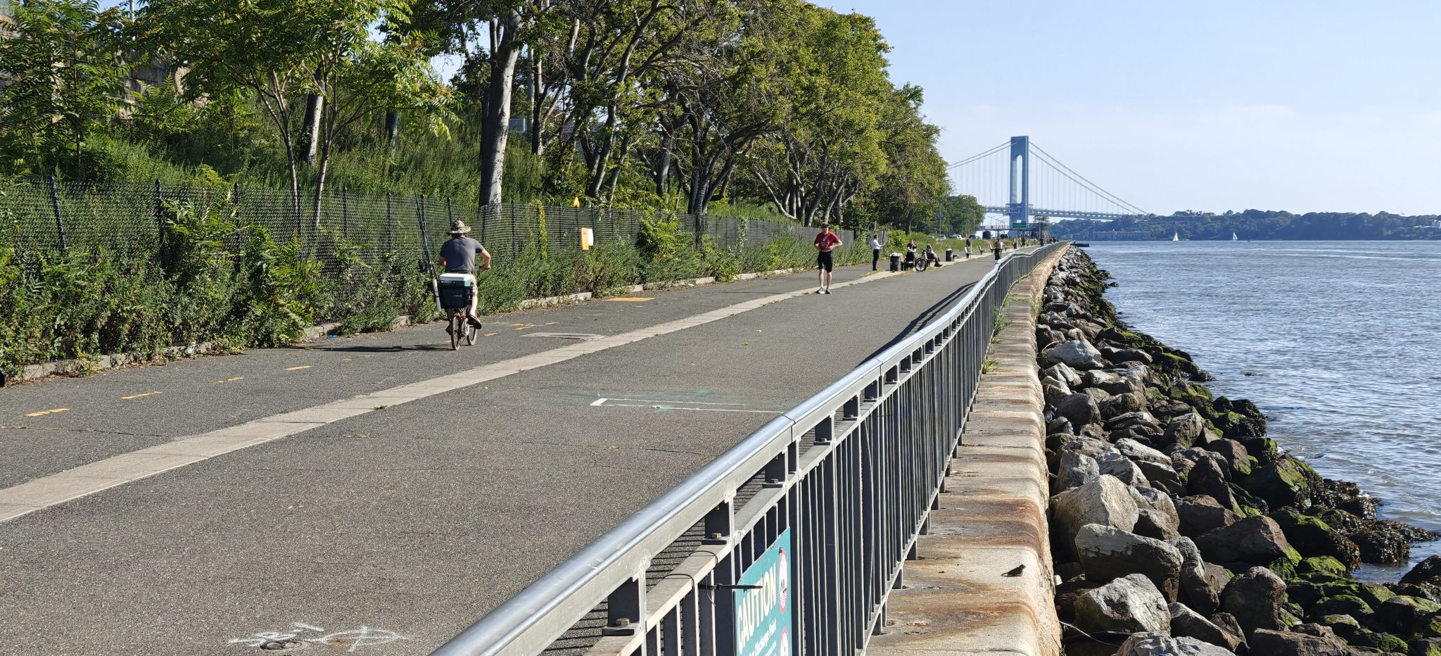 People cycling and running on a paved path by the water on a sunny day. - Shore Road Greenway