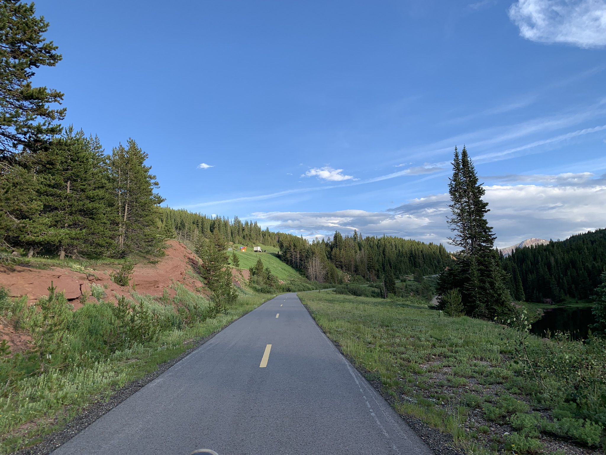Paved road winding through green pine forests and grassy hills under a blue sky. - Vail Pass