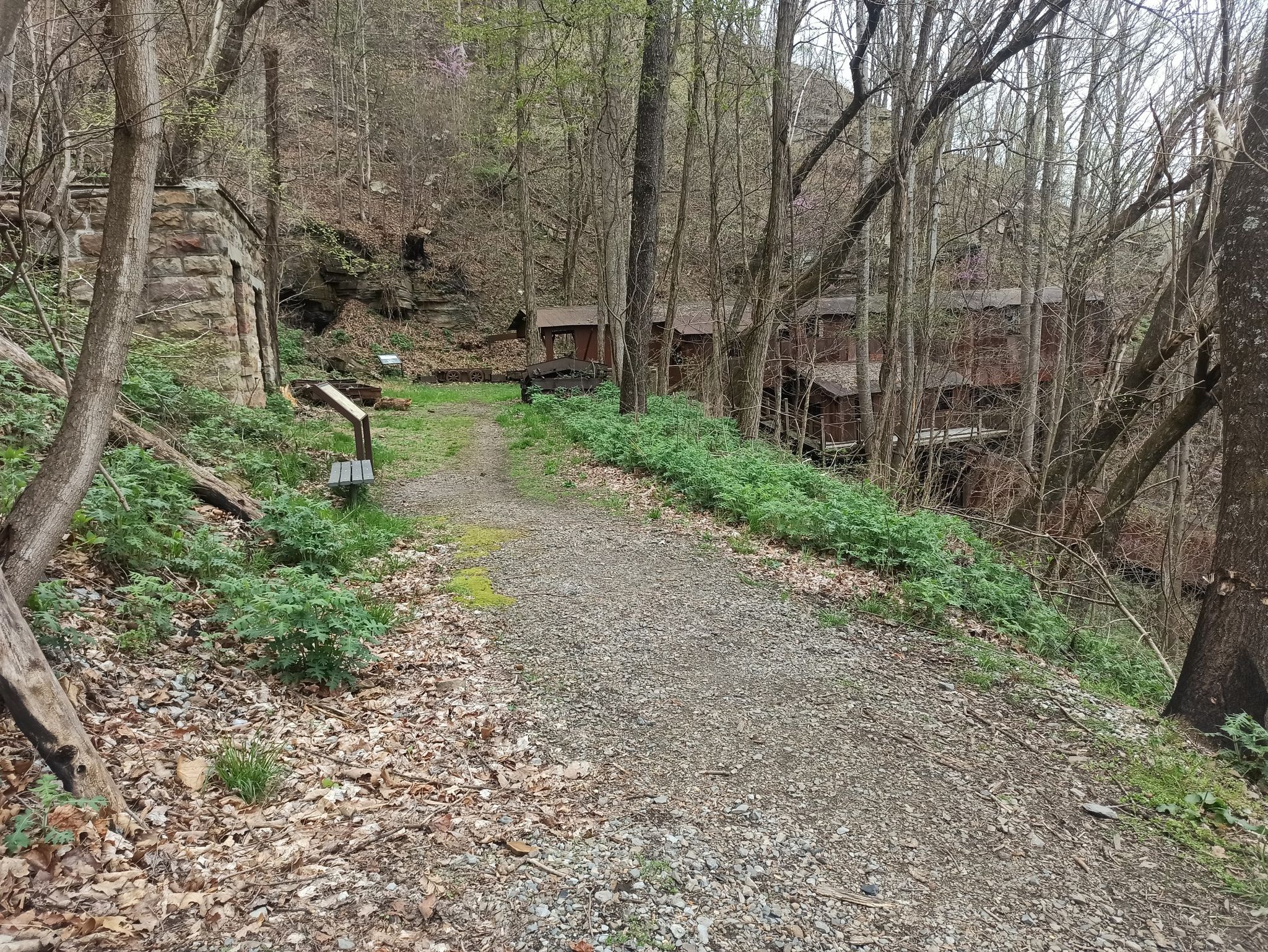 A gravel path winds past a stone wall and old industrial buildings in a forest. - Headhouse Trail to Nuttalburg Historic Site