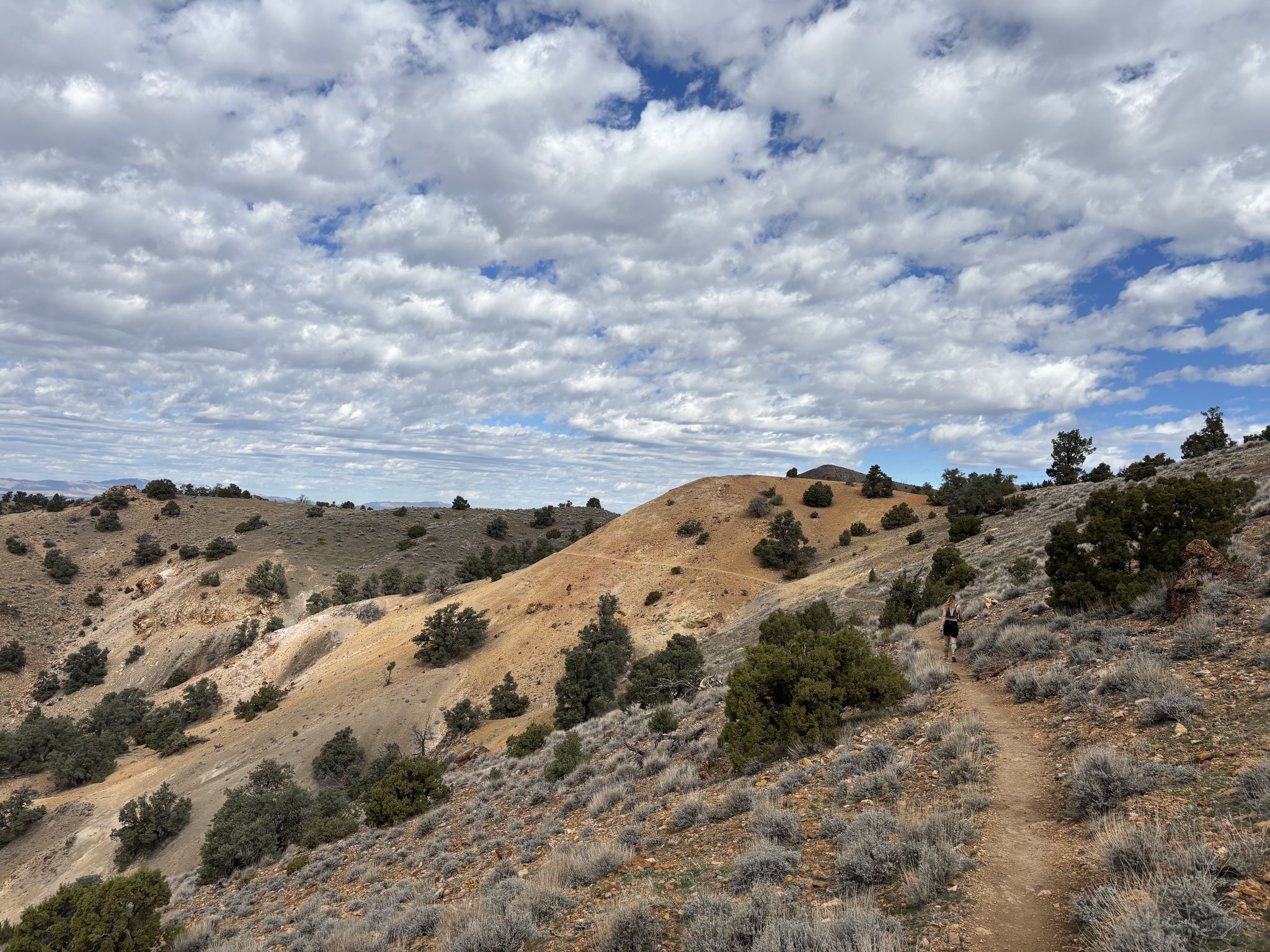 A person walks on a dirt trail winding through arid hills under a cloudy sky. - Hidden Valley Backcountry Loop