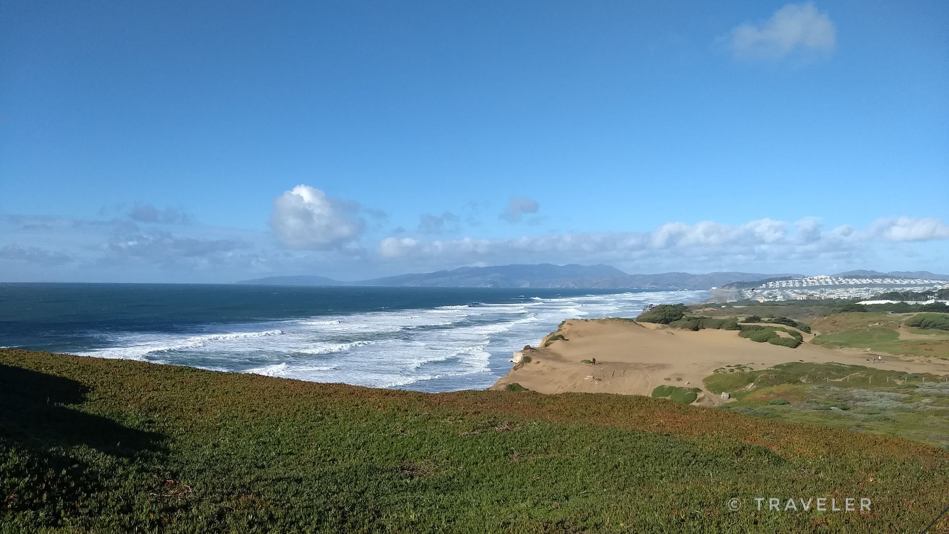 Ocean waves crashing on a sandy beach below a green cliff, clear sky. - Fort Funston Coastal Trail