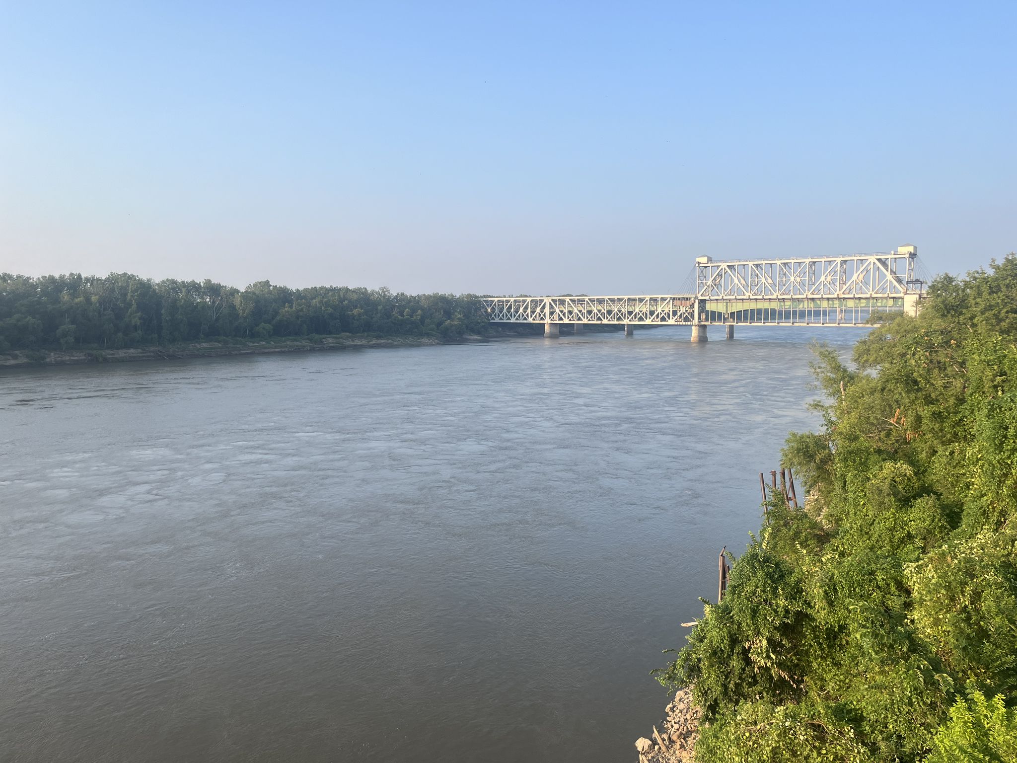 A long metal truss bridge spans a wide river under a clear sky. - Riverfront Heritage Trail