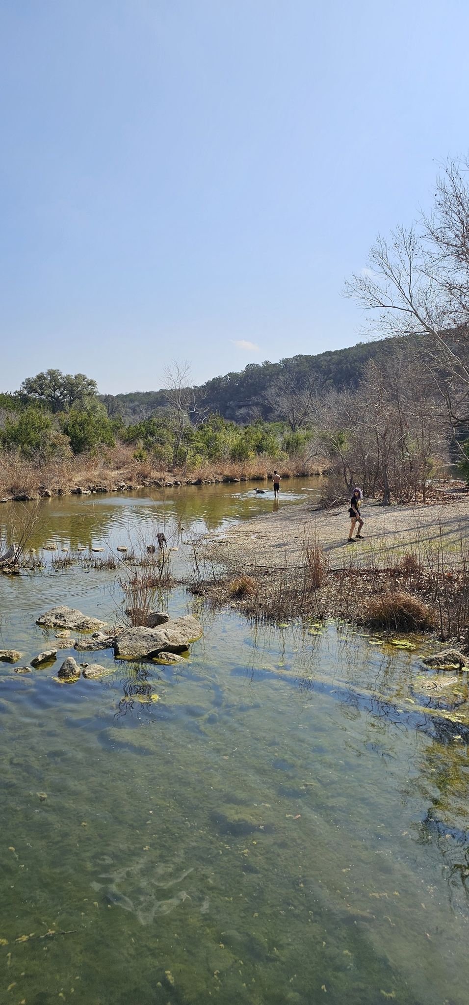 Sunny day by a clear river with people on the bank and distant hills. - Sculpture Falls and Hill of Life Falls Loop