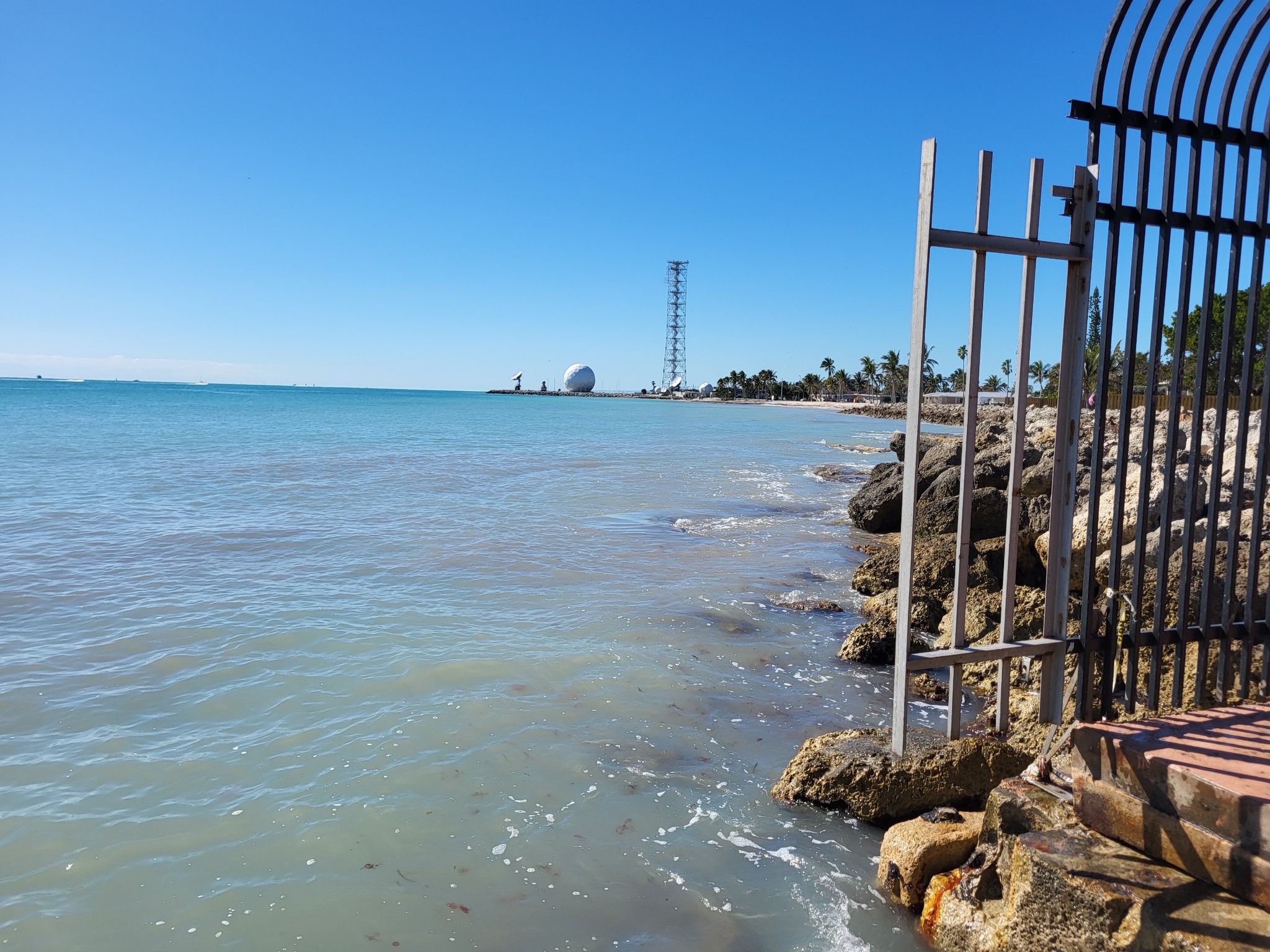 Rocky shoreline with a metal fence, calm ocean, and distant structures under clear sky. - Southernmost Point Buoy – Key West Lighthouse loop from Key West