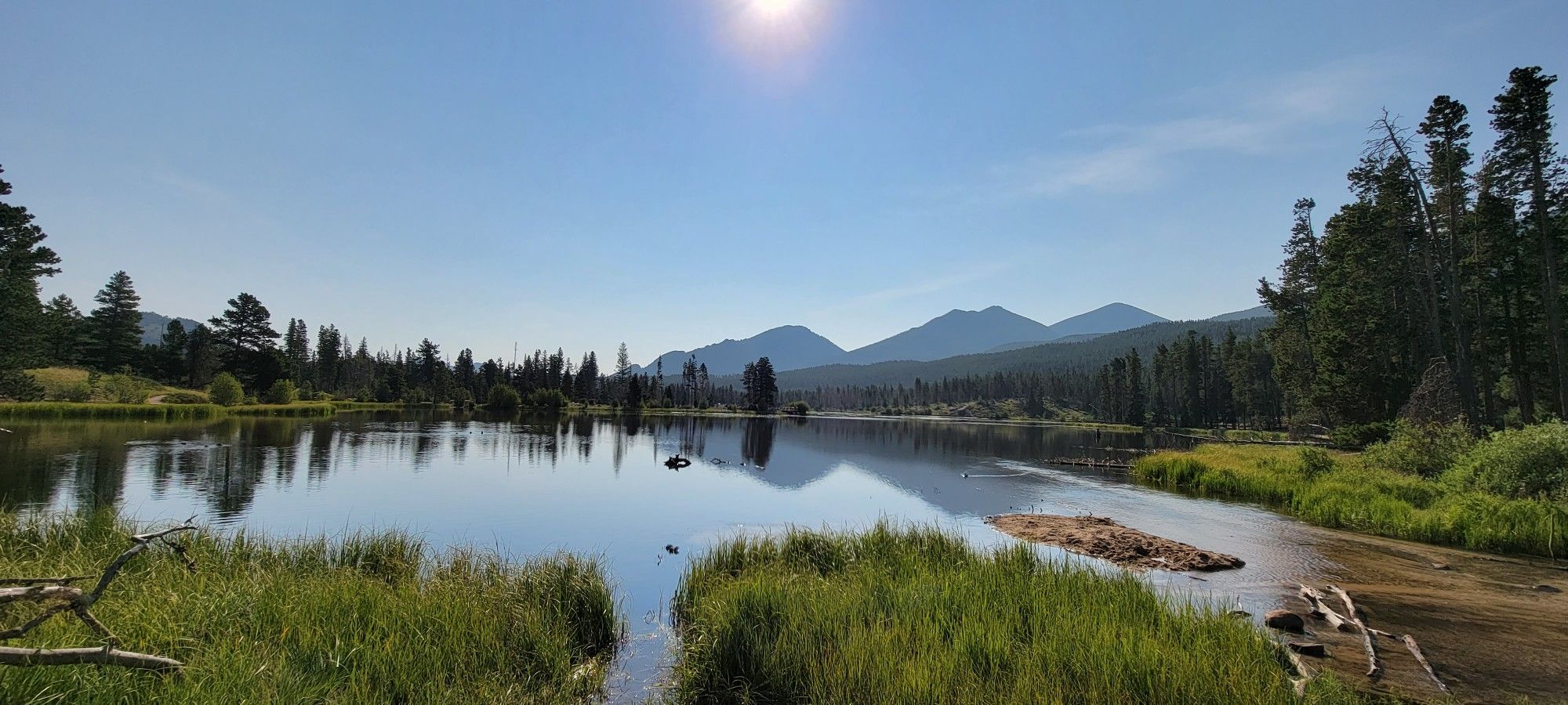 Bright sun over a calm lake reflecting distant mountains and pine trees. - Ride to Bear Lake