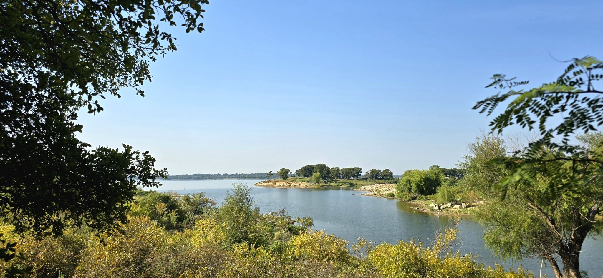 Panoramic view of a calm lake surrounded by green trees under a clear blue sky. - Northshore Trail
