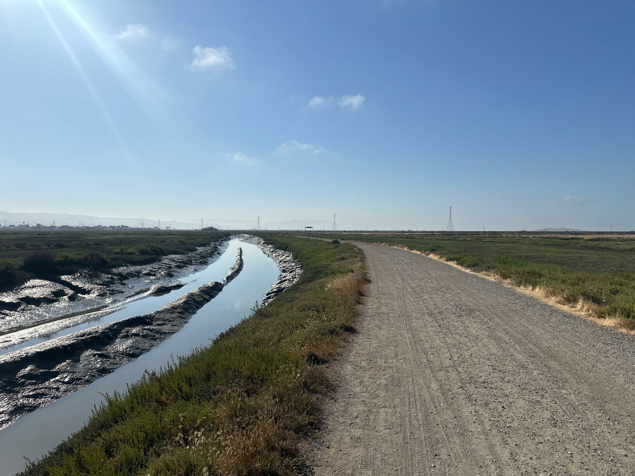 A dirt path next to a winding waterway through marshland on a sunny day. - San Francisco Bay Trail