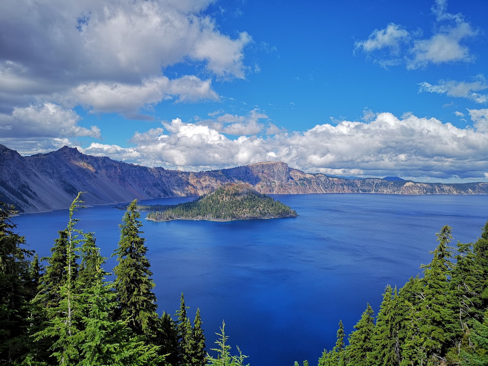 Vast blue lake with a forested island, surrounded by mountains under a bright sky. - Rim Village – Crater Lake National Park loop from Crater Lake Lodge