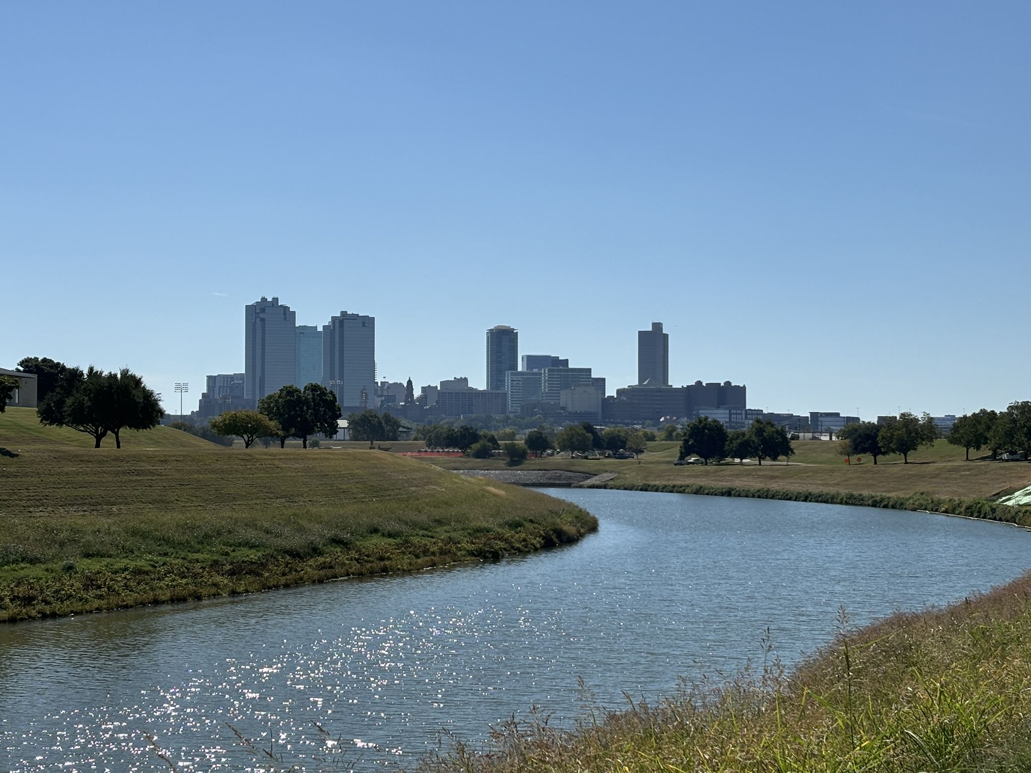 A city skyline across a winding river with grassy banks on a clear day. - Gateway Park West Loop