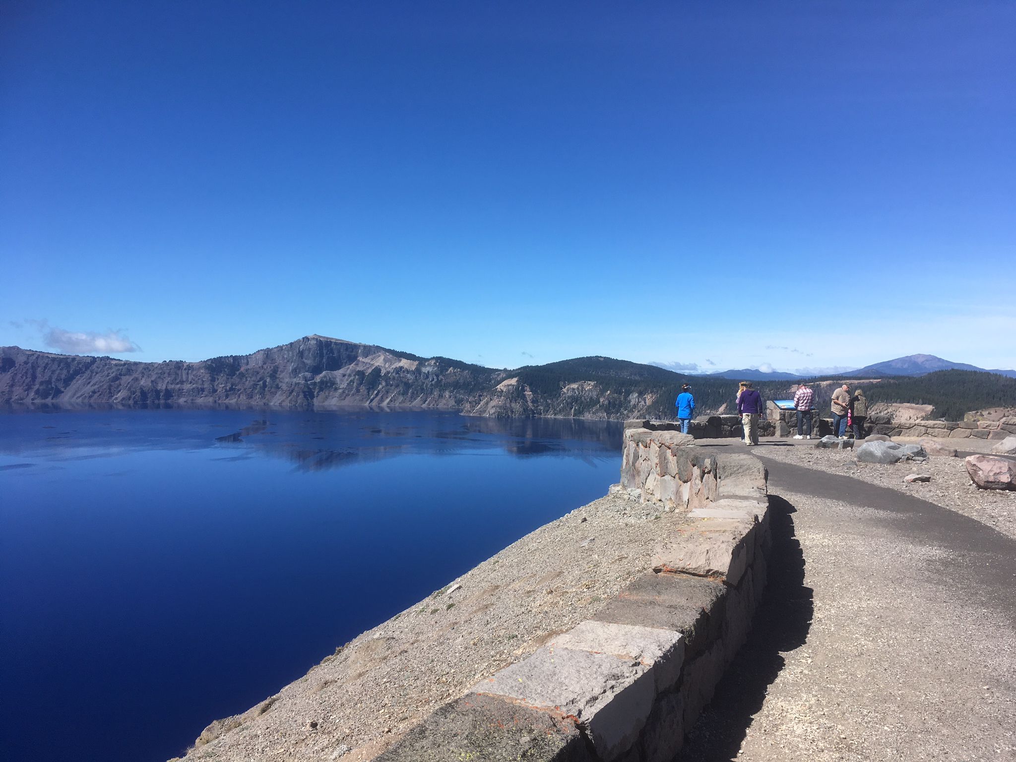 People on a stone overlook viewing a deep blue lake and distant mountains. - Skell Head to Cloudcap Viewpoint