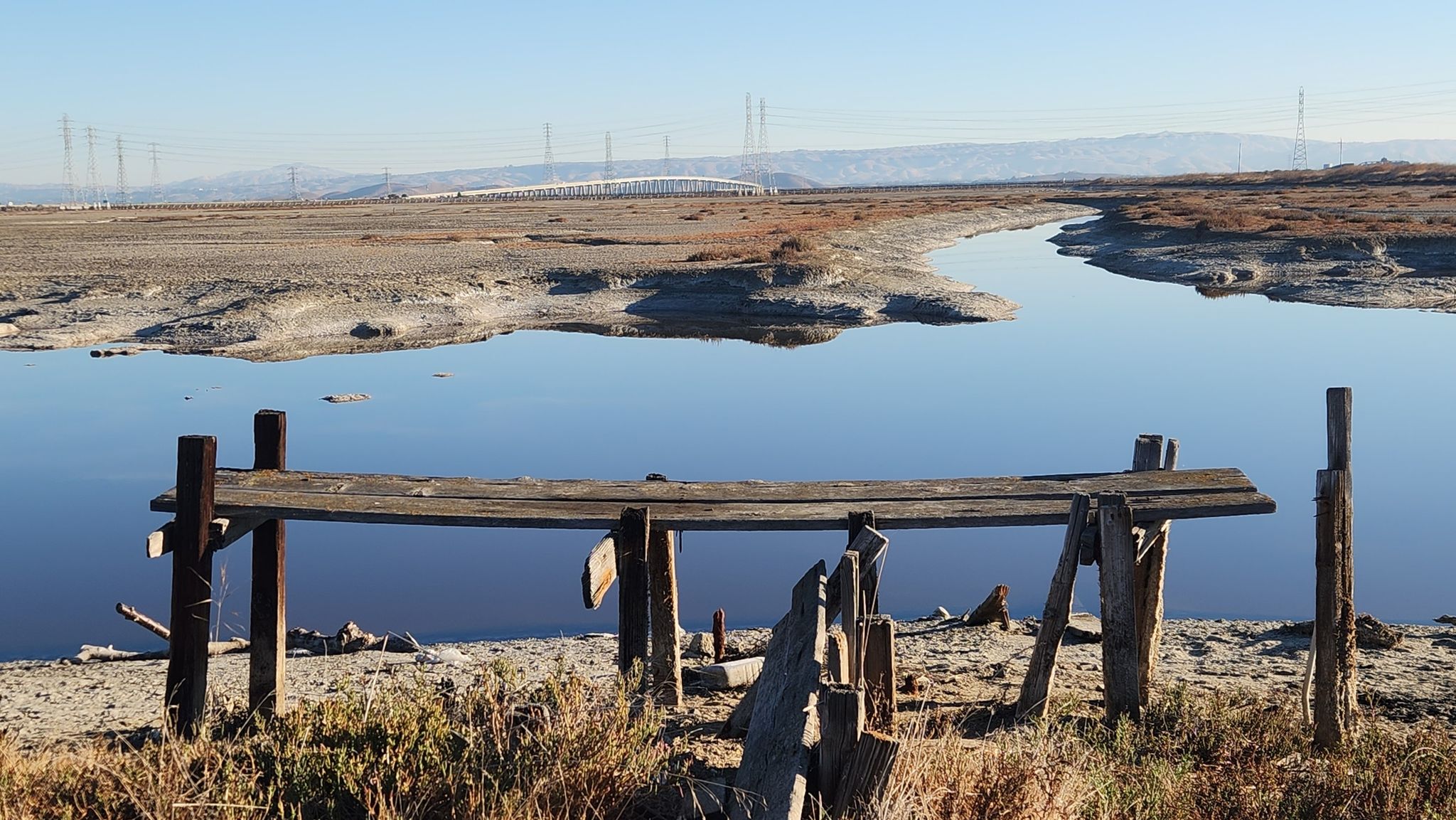 An old wooden structure stands by calm water with distant hills under clear sky. - San Francisco Bay Trail