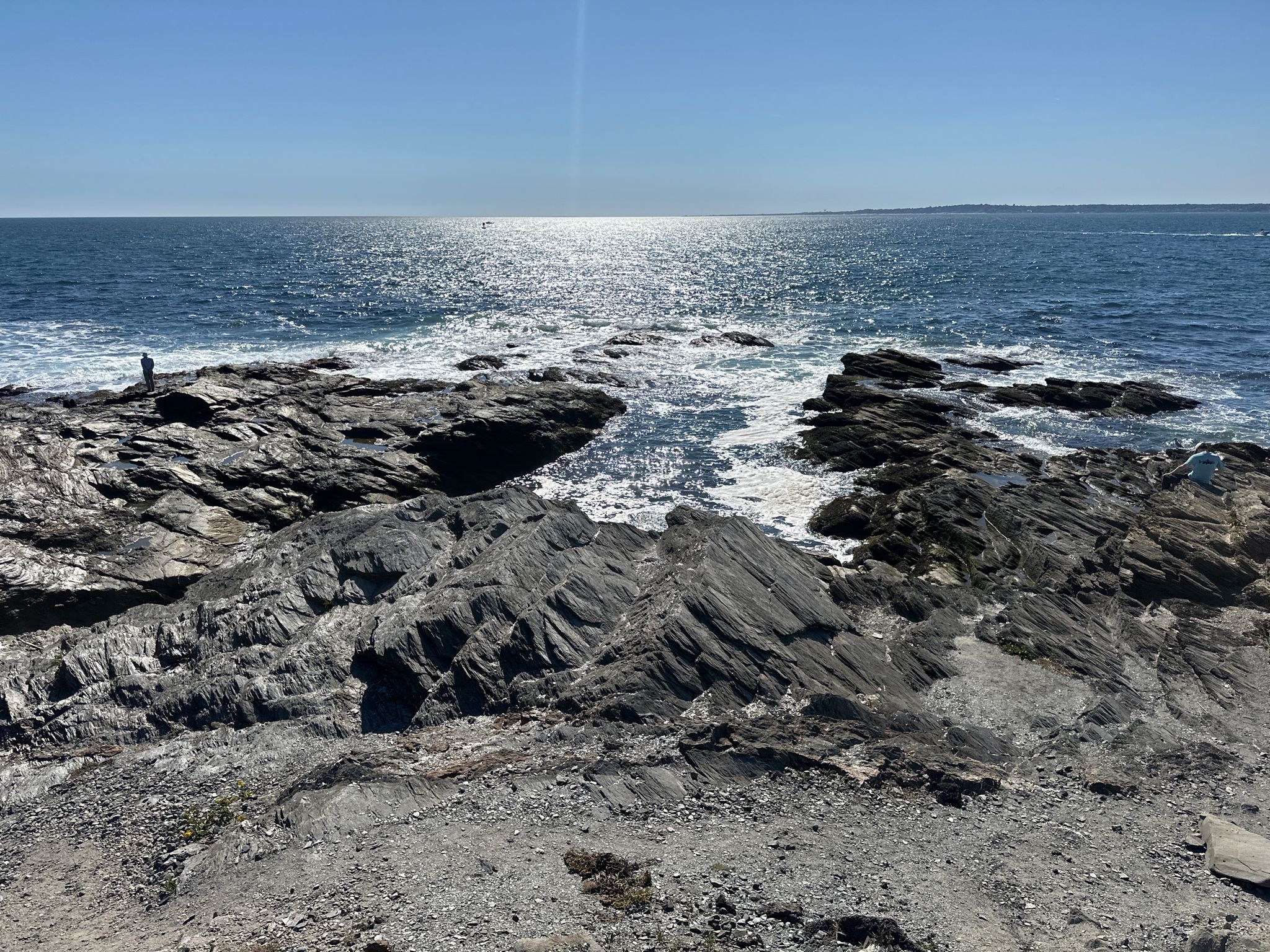 Rocky coastline with ocean waves, bright sun reflecting on the water, small person. - Beavertail Lighthouse Loop