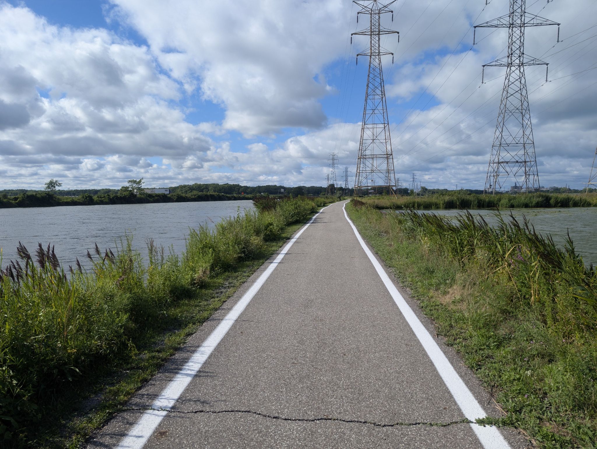 Paved path between two reedy lakes with power lines under a cloudy sky. - Wolf Lake Trail