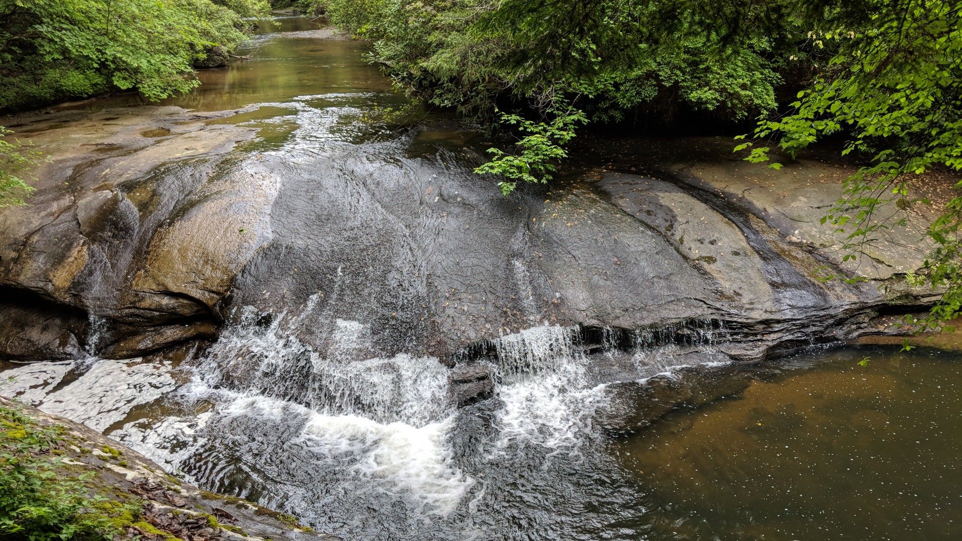 Wide, shallow waterfall flowing over smooth, dark rocks into a stream. - Eagle Falls Trail