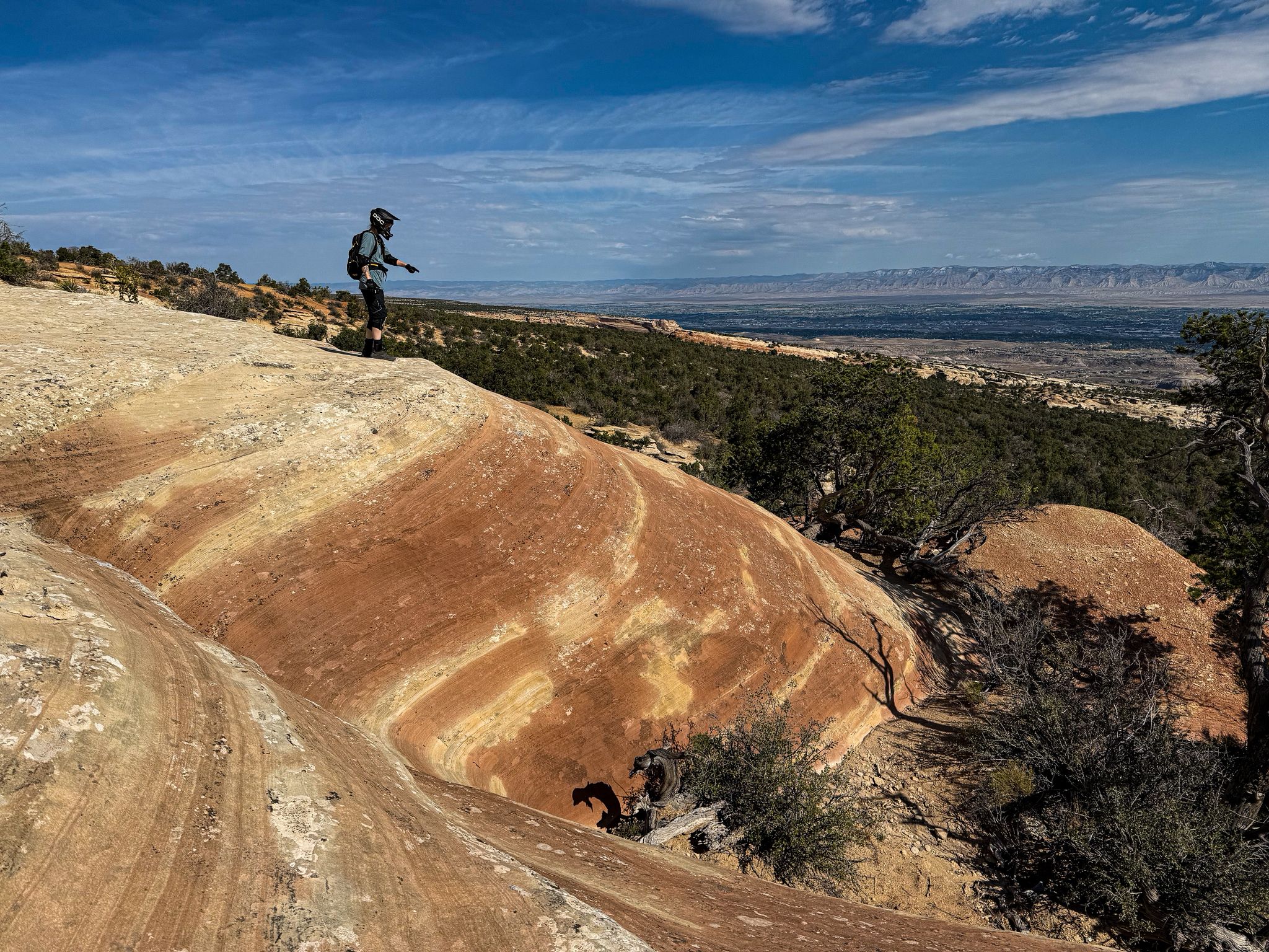 Biker on a rocky outcrop overlooking a vast valley under a bright sky. - The Ribbon Trail