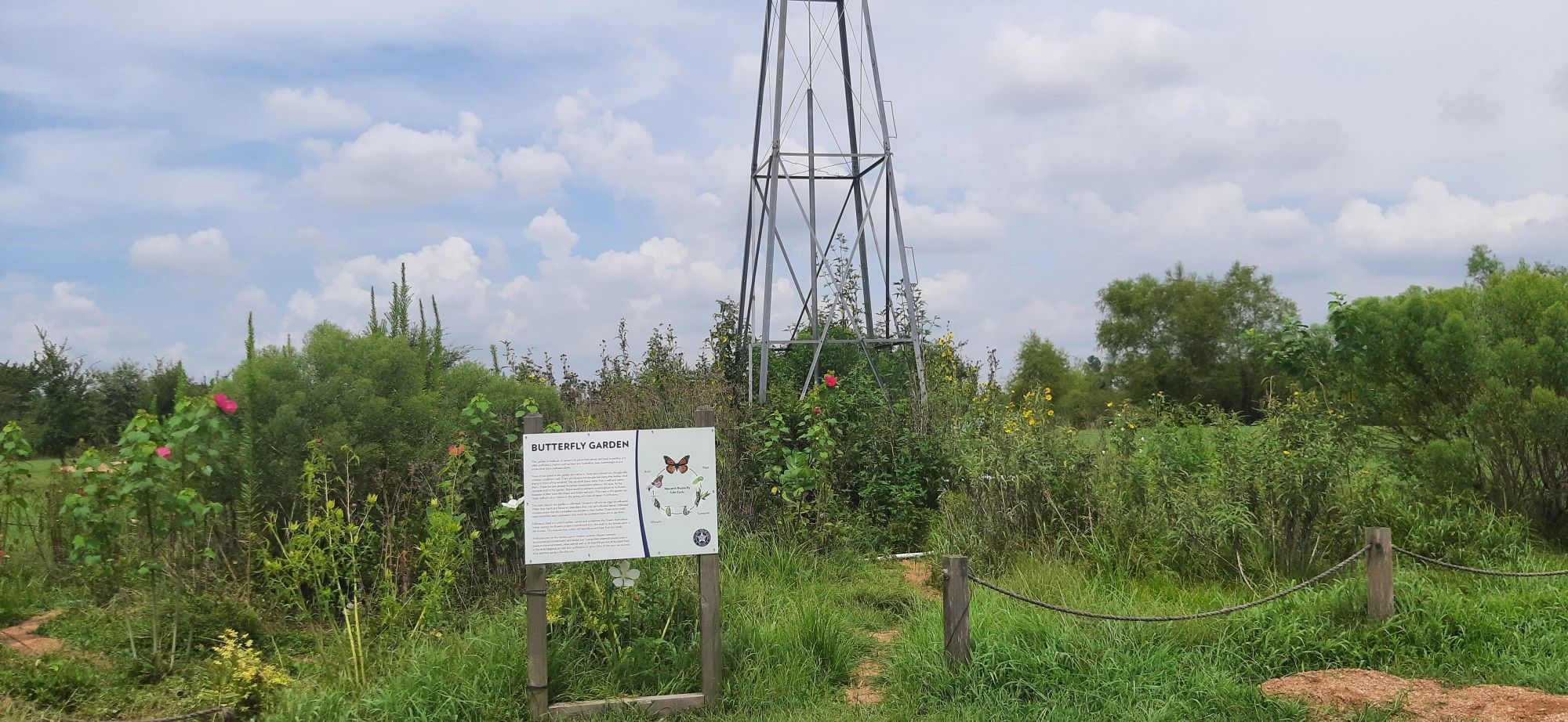 A tall windmill structure in a lush garden with a sign, under a cloudy sky. - Veterans Memorial Eagle – View of pond loop from Hightower High School