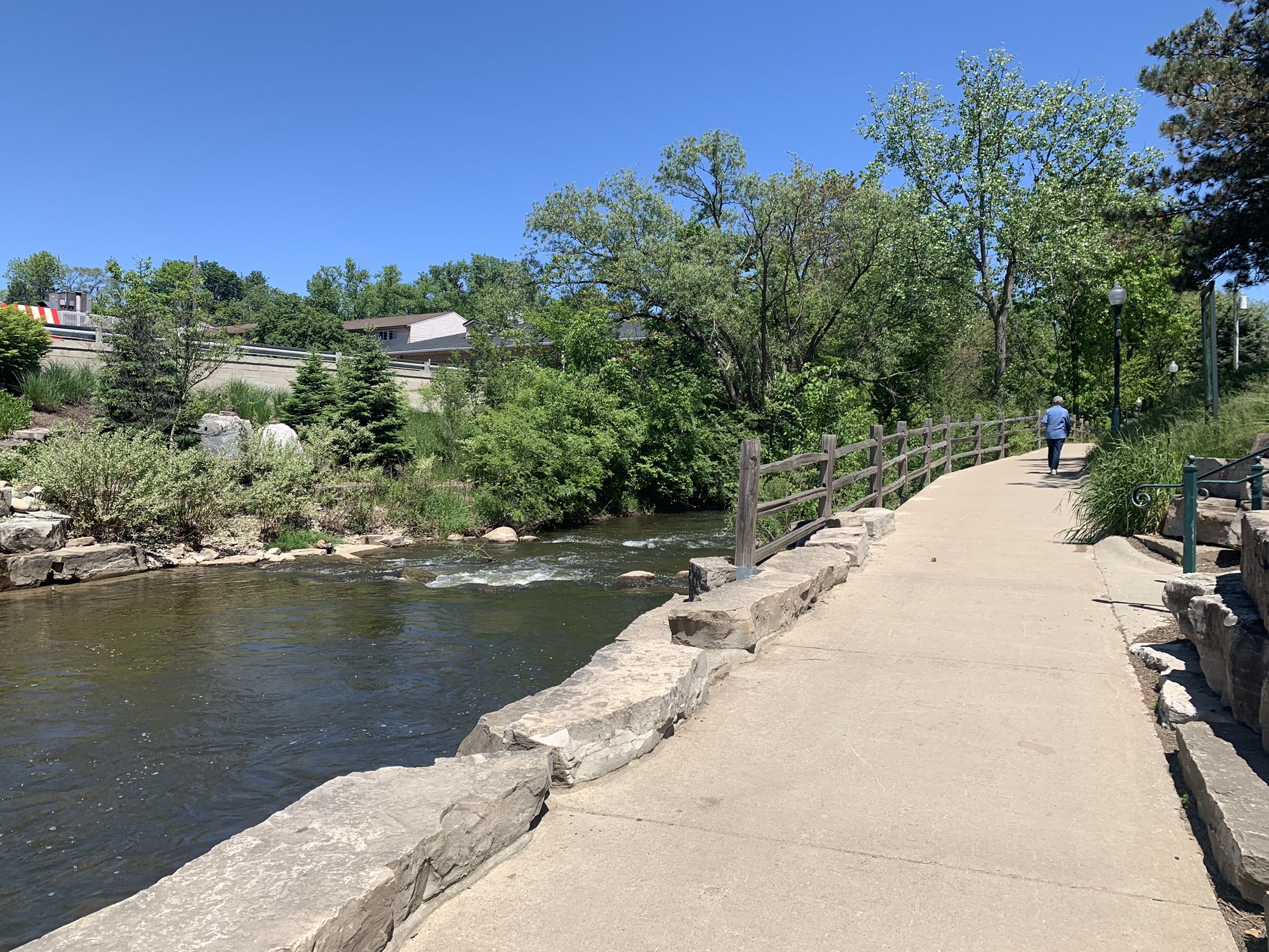 A person walks on a paved path beside a flowing river on a sunny day. - Rochester River Walk – Pines Trail loop from Rochester