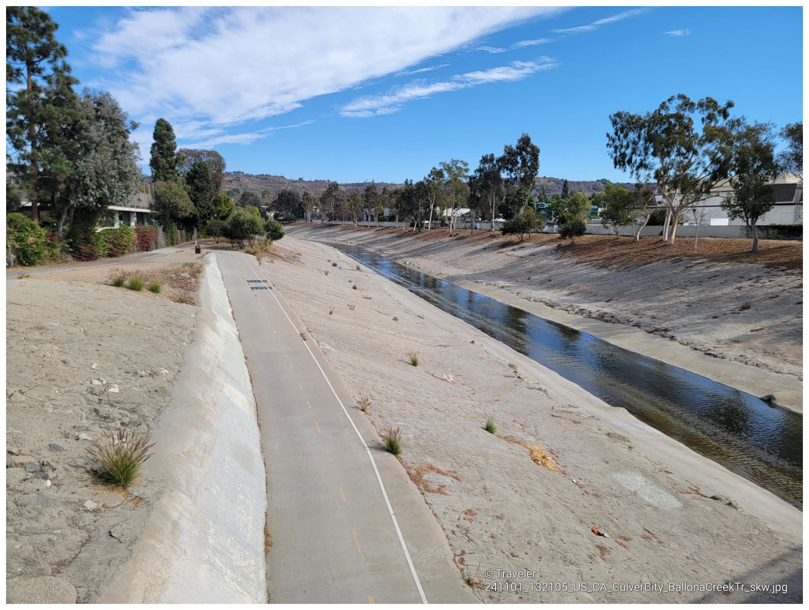 A concrete canal with a narrow path, surrounded by trees under a blue sky. - Rivers of the World Mural wall – Exposition Park Rose Garden loop from Downtown Inglewood