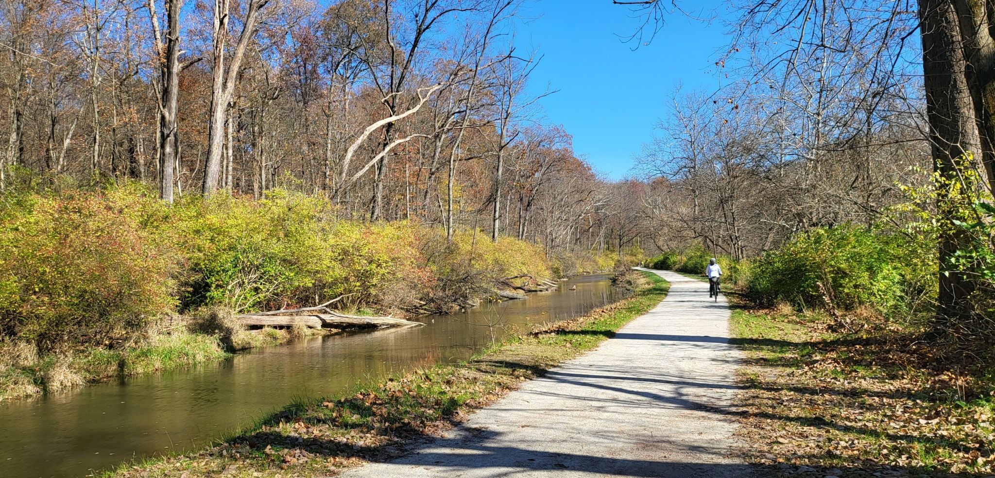 Dirt path next to a narrow canal, with a person cycling in the distance. - Ohio and Erie Canal Towpath: Rockside to Brecksville Station